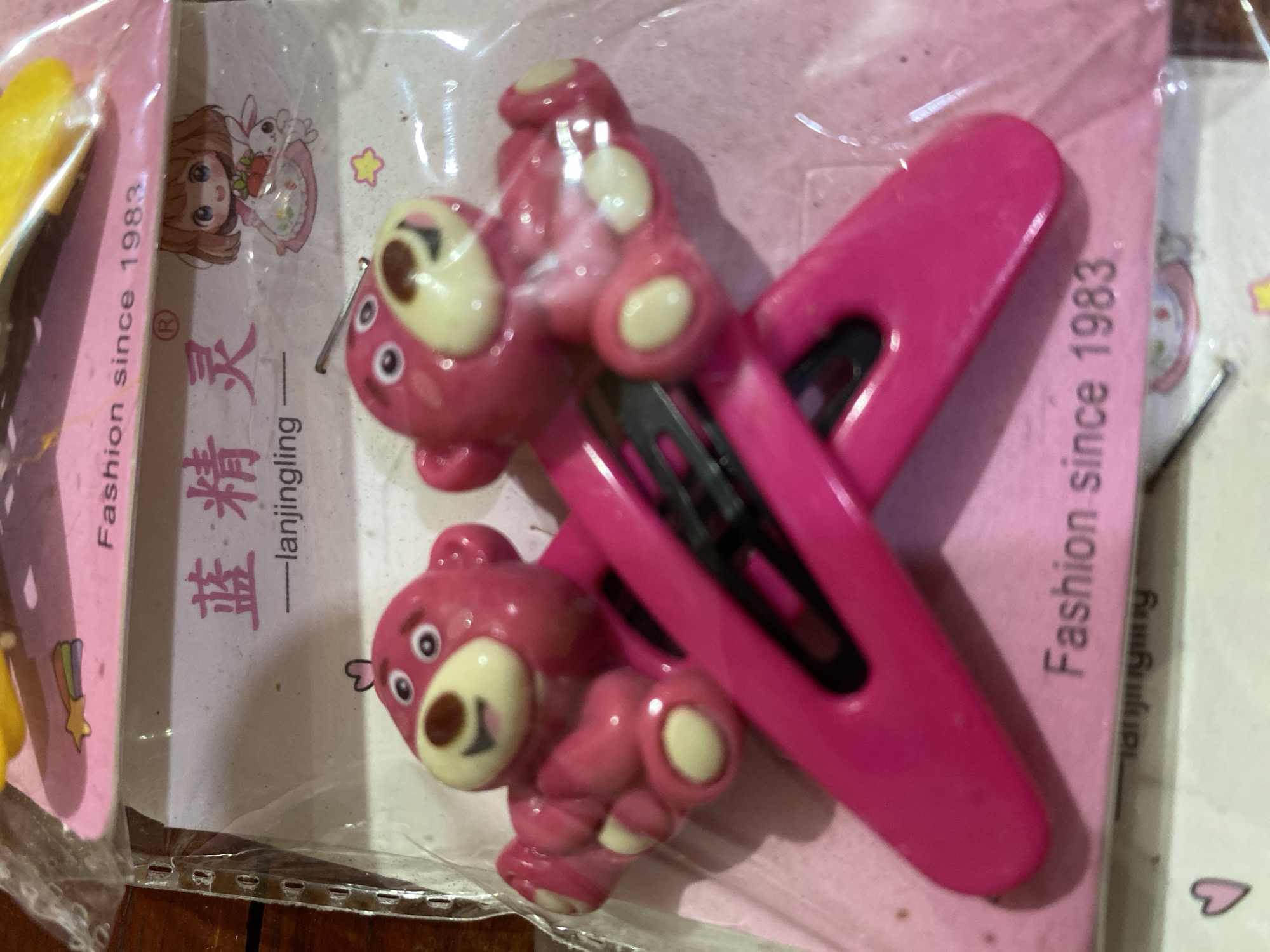 This is a close-up, eye-level, indoor shot of a package of pink bear-shaped hair clips. The package features a pink background with text and graphics. The text "Fashion since 1983" is visible in white. There is also Chinese text and an illustration of a girl with a bunny. The two hair clips are arranged diagonally across the center of the frame, with the bear shapes on top. The bears are pink with lighter pink accents and have black eyes and mouths. The hair clips themselves are a bright pink color. The lighting is slightly diffused, and there are some reflections on the plastic packaging. The image appears to be taken in Ta', Indonesia, as indicated by the user prompt.