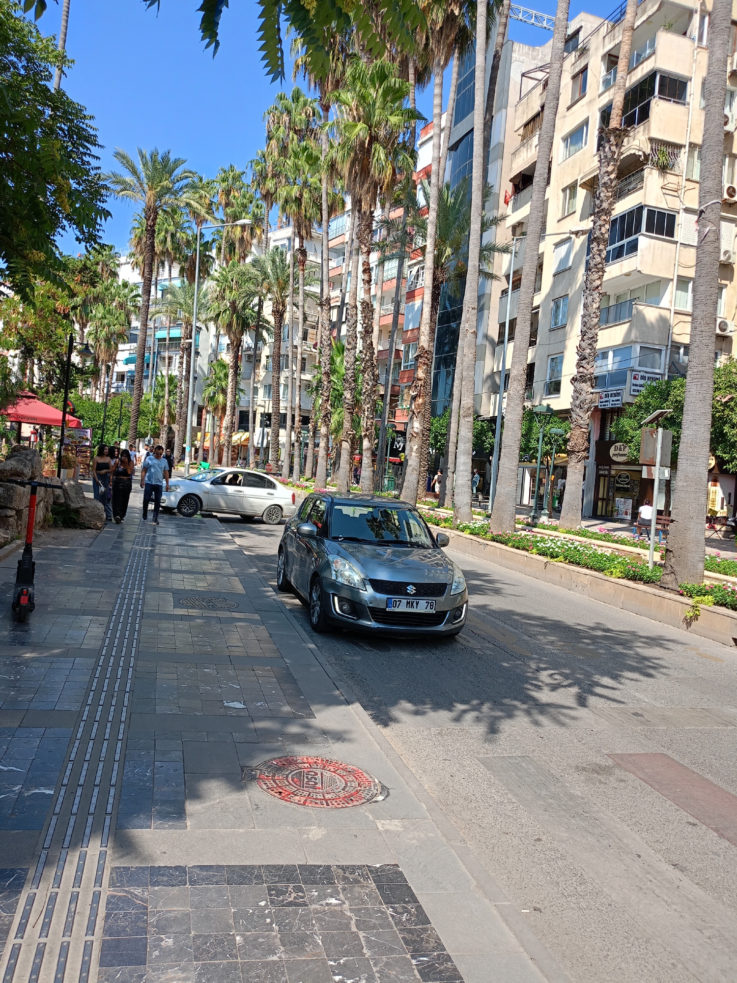 A street scene in Antalya, Turkey, features a road with two cars, one silver sedan and one gray hatchback, driving on it. The hatchback, a Suzuki, has a license plate reading "07 MKY 78". A sidewalk runs alongside the road, paved with dark and light grey tiles, and features tactile paving for the visually impaired. An electric scooter is parked on the sidewalk. Several palm trees line the sidewalk, casting shadows on the pavement. In the background, a multi-story building with balconies and windows stands tall. A few people are walking on the sidewalk. The sky is clear and blue.