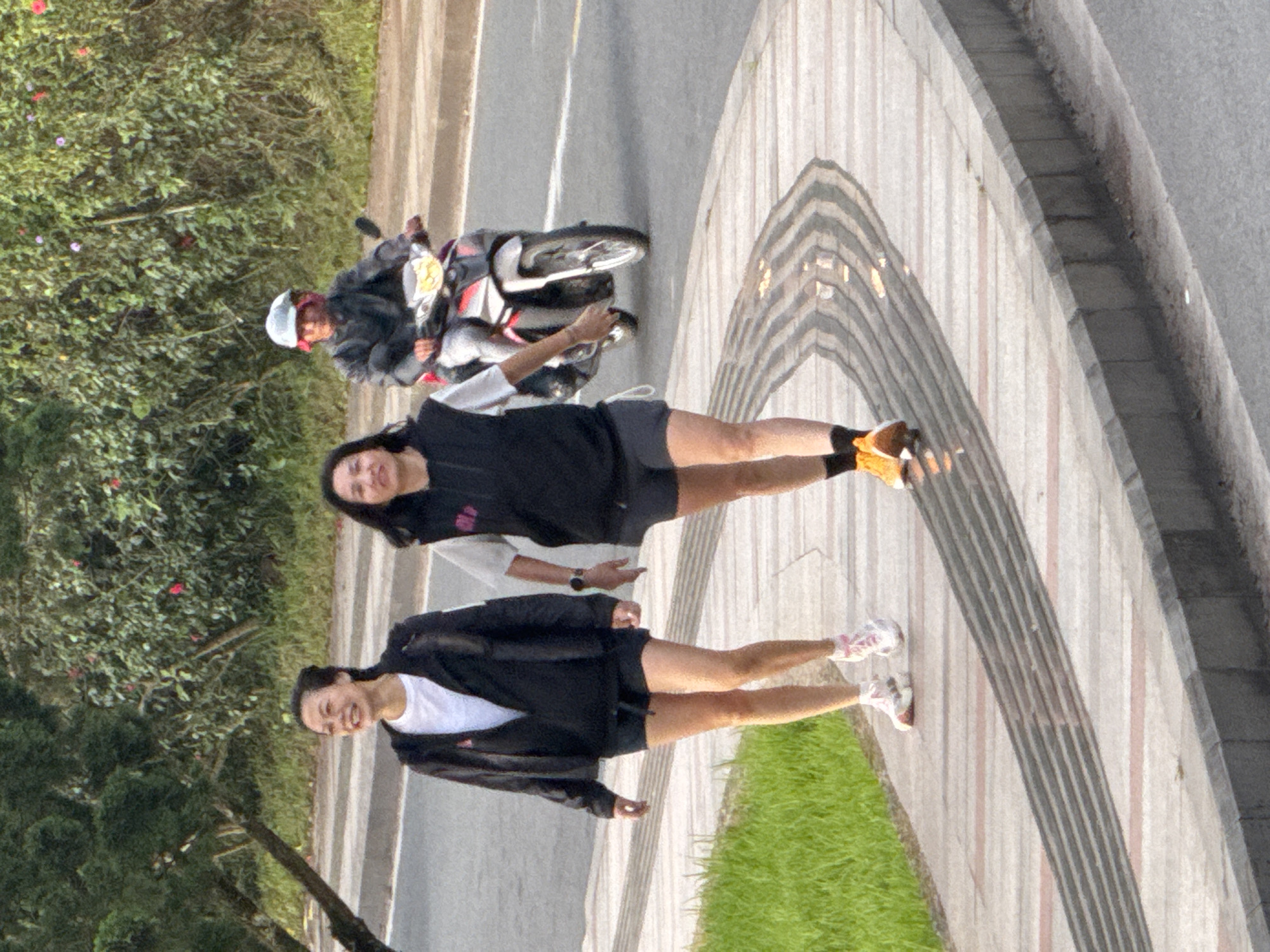 The image depicts an outdoor scene in Phường Xuân Hương - Đà Lạt, Vietnam, featuring two women walking on a sidewalk parallel to a road where a motorcyclist is riding.
In the foreground, two East Asian women are walking from left to right on a light-colored paved sidewalk. The woman on the left is smiling with her mouth open, revealing teeth. She wears a black long-sleeved jacket over a white t-shirt, black shorts, and white and pink athletic shoes with white socks. The woman on the right has a neutral or slightly smiling expression. She wears a black sleeveless vest over a light-colored t-shirt, dark shorts, and bright yellow/orange athletic shoes with black socks. A dark-faced watch is visible on her left wrist. The sidewalk features a section with parallel dark grey lines, possibly tactile paving, and is bordered by a raised concrete curb on the right and green grass on the left.
In the mid-ground, on the asphalt road to the right of the women, a person is operating a silver and red scooter-style motorcycle. The operator wears a light grey helmet, a dark jacket, and a dark red scarf or cloth around the neck. The motorcycle's headlight is illuminated.
The background consists of dense green foliage and trees, with some small red and pink flowers visible within the greenery. The lighting suggests natural daylight, possibly morning or late afternoon.