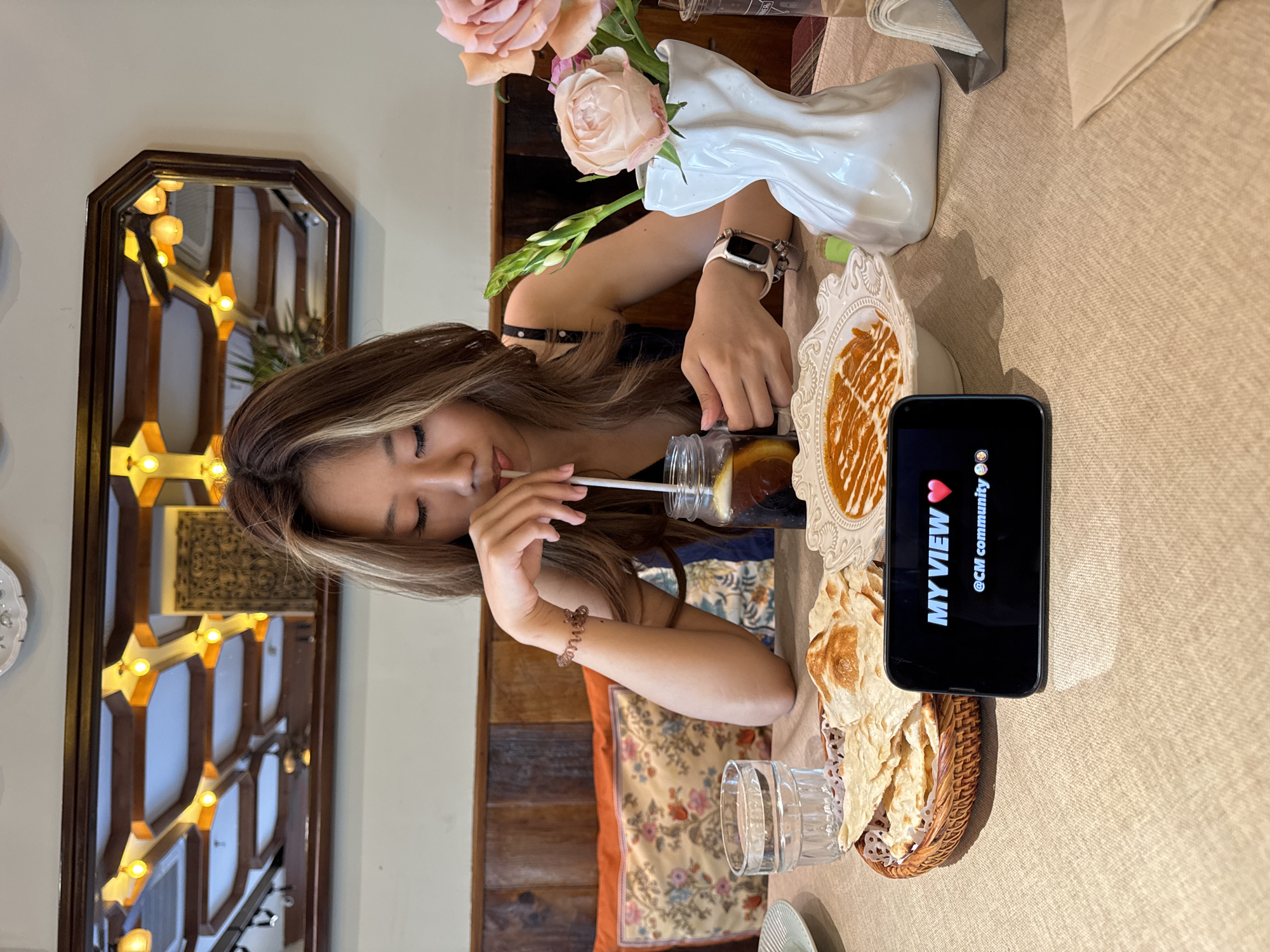 A woman is seated at a table, drinking from a glass with a straw. A cell phone stands upright on the table, displaying "MY VIEW" and "@CM community." There is food on plates and in a basket next to the phone, and a vase of flowers sits behind the food. The woman has long brown hair and is wearing a black top. A mirror with decorative lights is visible in the background. The scene appears to be inside a restaurant or cafe. This image was taken in Yuzhong District, China.