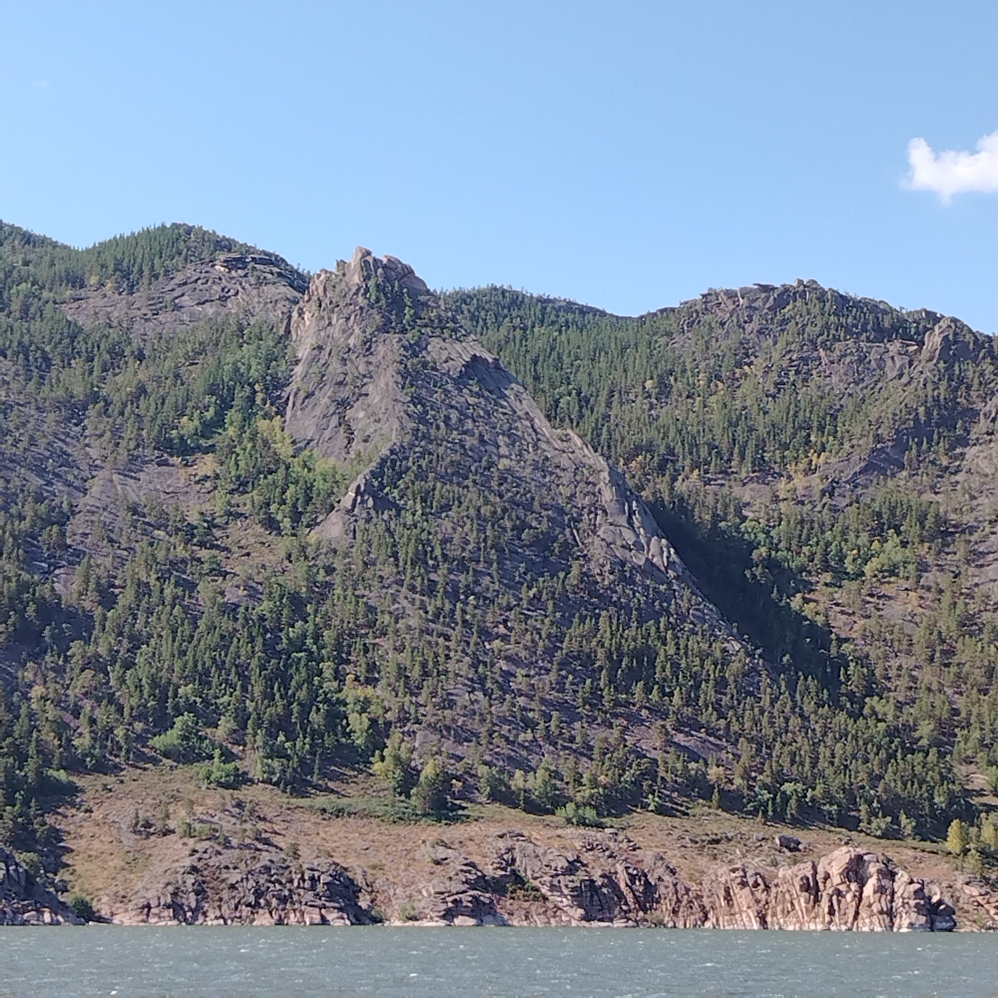 This is a wide-angle, landscape photograph taken on a clear, sunny day from the perspective of a body of water. The image features a large, rugged mountain range covered in dense green pine trees. The mountains are characterized by steep, rocky slopes and jagged peaks. In the foreground, the dark, choppy water reflects the blue sky. The shoreline is rocky, with exposed rock formations meeting the water. To the right of the frame, a small, wispy white cloud is visible in the sky. The scene is natural and undeveloped, suggesting a remote or natural park area within or near the city of Торайғыров, Kazakhstan, known for its mountainous terrain. There are no people or animals visible in the image.
