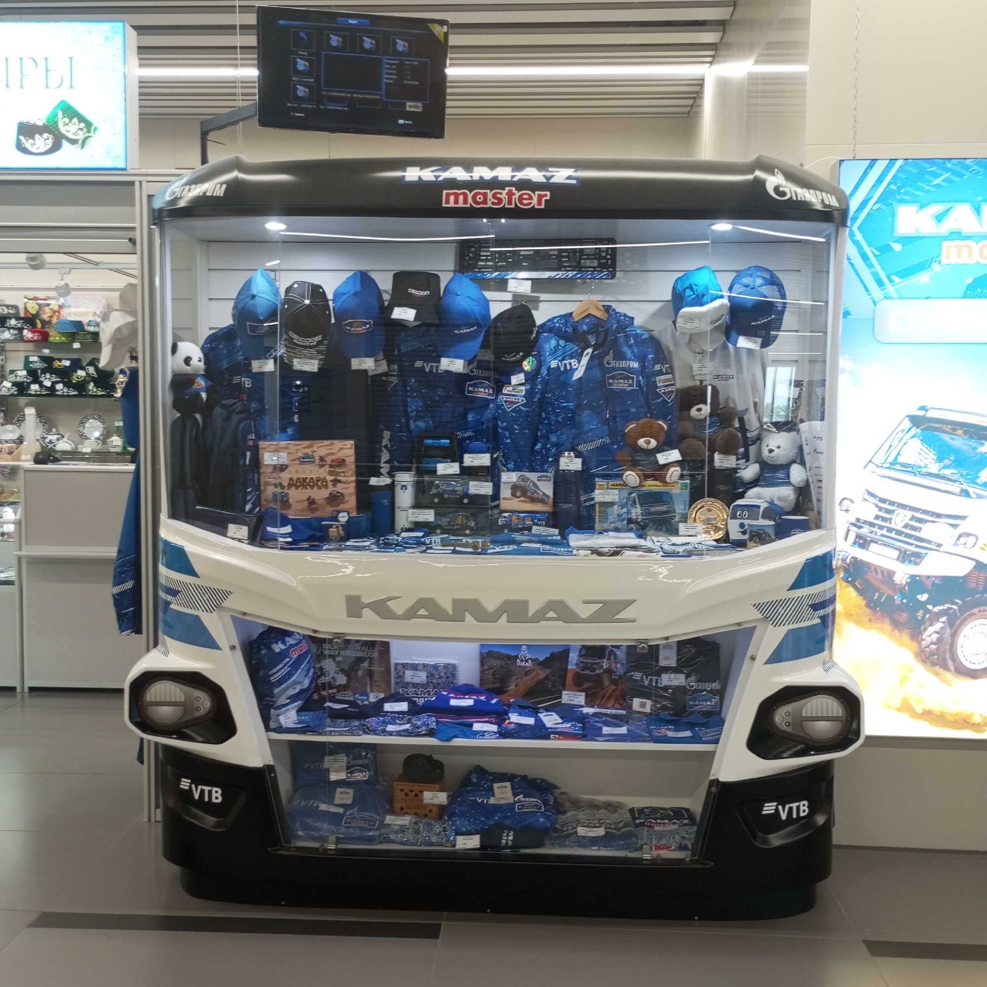 The image shows a store display in Naberezhnye Chelny, Russia, featuring merchandise related to KAMAZ trucks. The display is designed to resemble the front of a KAMAZ truck, complete with headlights and a grille. Inside the display, various items such as hats, jackets, t-shirts, and model trucks are arranged on shelves. The merchandise is predominantly blue and white, the corporate colors of KAMAZ. Logos for KAMAZ and VTB Bank are visible on the products and the display itself. In the background, there are other store shelves with additional merchandise, and a large poster of a KAMAZ truck. A television screen is mounted above the display, showing what appears to be a dashboard or informational graphic. The environment is well-lit, suggesting an indoor retail space. There are no people visible in the image.