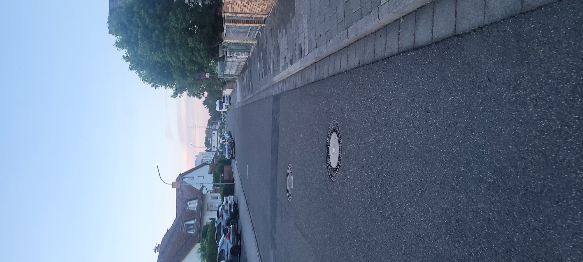 The image depicts a quiet residential street in Heusenstamm, Germany, likely during twilight hours, evidenced by the light blue sky with a faint pinkish tint and an illuminated street lamp. The perspective is from the center of the asphalt road, looking down the street.
On the left side of the road, a row of two-story houses with pitched, dark-tiled roofs and light-colored facades are visible, some featuring dormer windows. Green hedges and bushes line the front of these properties. Several cars are parked along the left curb, including a dark grey SUV and a silver sedan in the immediate foreground, and a dark blue Volkswagen Golf further down the street. A modern street lamp with an active LED light source is centrally positioned on the left sidewalk.
The right side of the scene features a grey paved sidewalk with a distinct darker curb. Beyond the sidewalk, a brick fence with a metal gate is visible in the mid-ground, followed by a lighter-colored, weathered fence partially overgrown with greenery. Mature green trees line this side of the street, and a red circular prohibitory sign is affixed to a pole or tree. Further down the street on the right, a white camper van and another white vehicle are parked.
The dark grey asphalt road surface features two prominent circular metal manhole covers in the foreground. In the distance, a utility pole with power lines is discernible. No individuals or active interactions are present in the scene.
