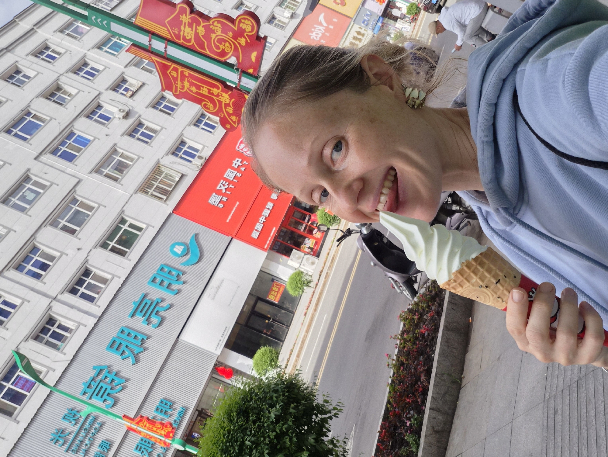 A woman is taking a selfie while holding an ice cream cone and sticking her tongue out. She is wearing a light blue hoodie and a decorative earring. Behind her, a building with Chinese characters on its facade and windows is visible. The building appears to be in the city of Mishan, China, as indicated by the signage. The street in front of the building has a sidewalk, a road with a motorcycle, and a person sitting on a stool in the background. Red flowers and green bushes line the sidewalk. The overall scene suggests an urban environment.
