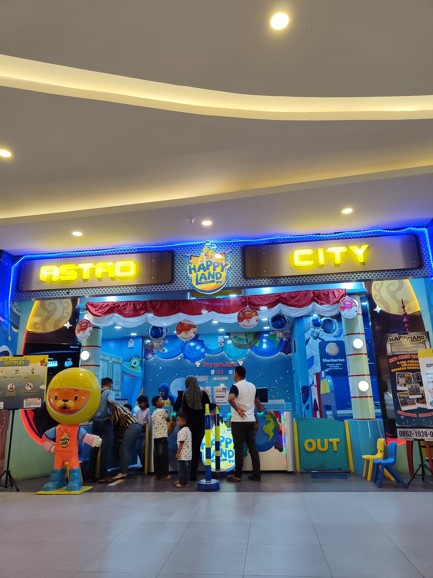 This image captures the entrance to "Happy Land Astro City" in Sempusari, Indonesia. The facade is brightly lit with blue neon lights outlining the words "ASTRO" and "CITY" in large, yellow, illuminated lettering. A stylized castle graphic with the words "HAPPY LAND" is also visible. Red and white bunting hangs above the entrance, and the interior appears to be a themed play area with space-related decor, including planets, rockets, and astronaut figures.

In the foreground, a large mascot statue of a cartoon lion in an orange astronaut suit stands to the left. Several people, including adults and children, are gathered near the entrance, some appearing to be waiting or entering the facility. One person is standing at a counter or booth to the right of the entrance. The floor is tiled, and yellow and blue children's chairs are placed near the right side of the entrance. Posters and signage are visible on the right, including one with a phone number. The overall environment suggests an indoor entertainment venue for families.