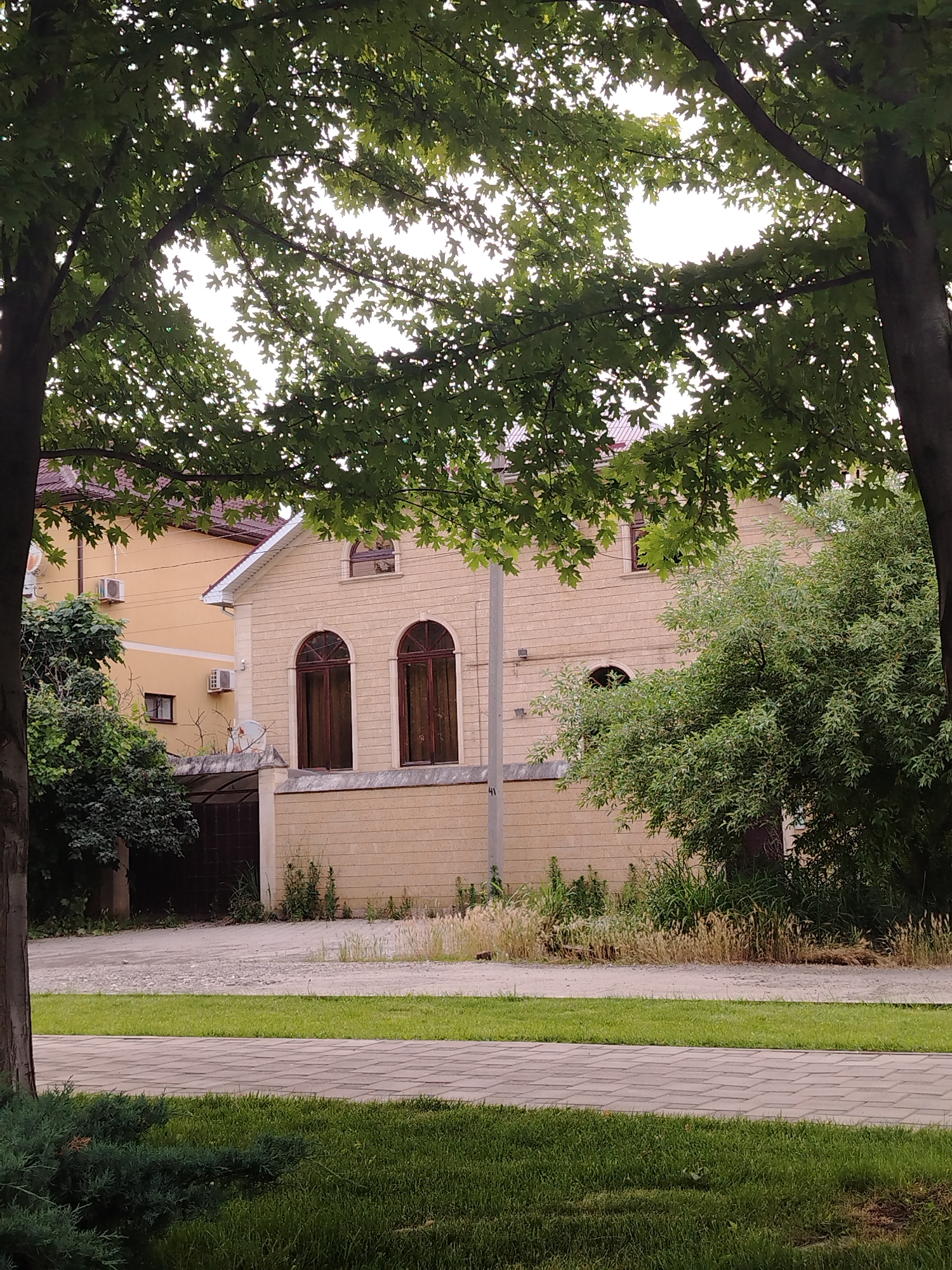 The image depicts a two-story building with a light beige brick facade and arched windows, possibly in Krasnodar, Russia. The building's roof is a dark red tile. To the left of the main building, a yellow building is visible with air conditioning units attached to its exterior. A low wall made of the same beige brick runs in front of the buildings, topped with a dark metal fence. Green trees with dense foliage are prominent in the foreground and around the buildings. The ground in front of the wall is paved with grey interlocking stones, and beyond that, a strip of green grass is visible, followed by a gravel driveway. The sky is overcast, suggesting it is daytime. There are no people or animals visible in the image.
