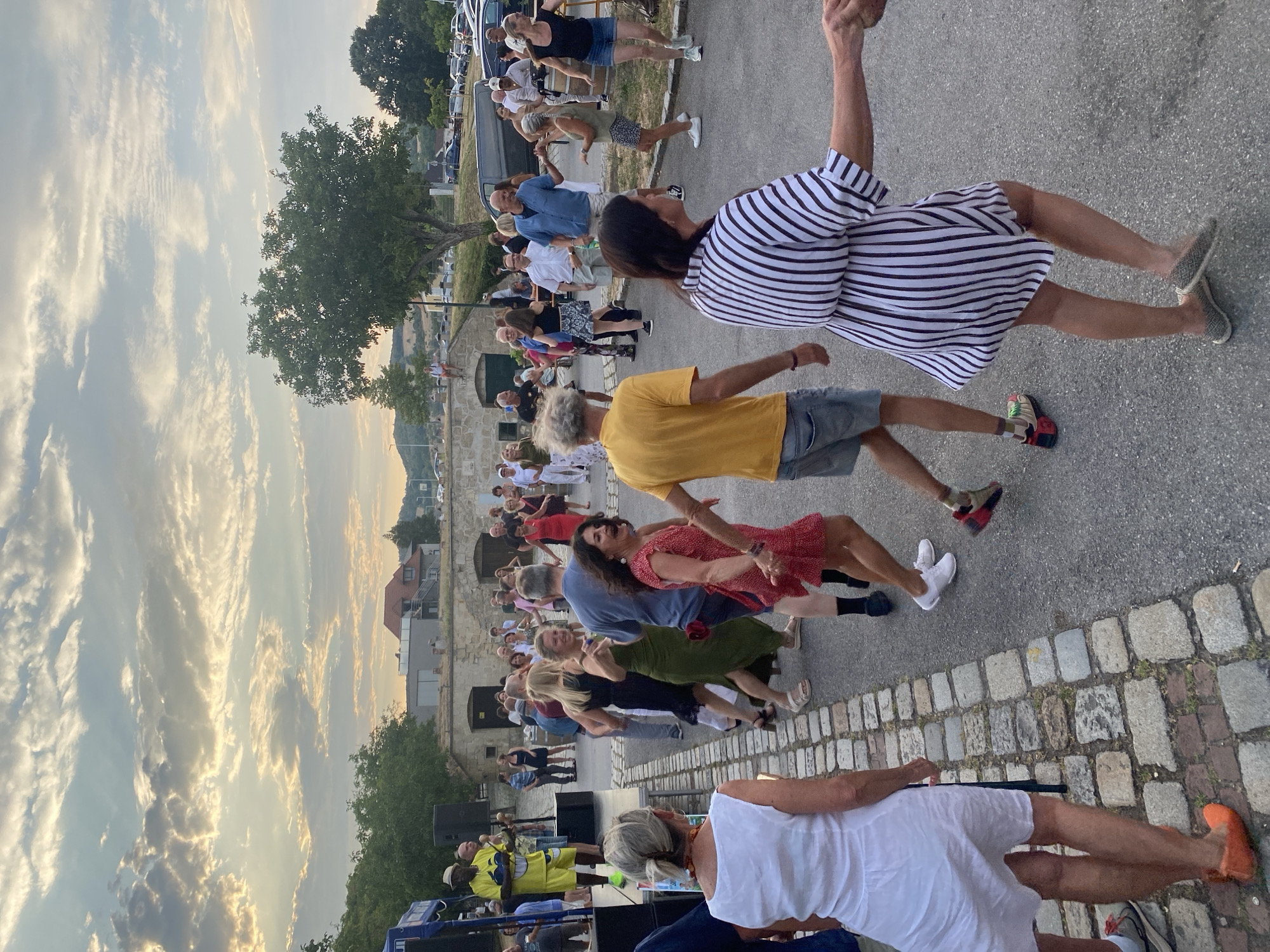 In Purbach, Austria, a group of people are dancing in a circle outdoors. The sky above is filled with dramatic clouds, illuminated by the setting sun. The dancing is taking place on a paved area next to a stone building, possibly a historic fortification, with a tree and further buildings visible in the background. Many of the individuals are dressed in casual summer attire, and some are holding hands as they move in unison. A stage with sound equipment is set up to the left, suggesting a musical performance is accompanying the dancing. The overall atmosphere appears to be festive and social.
