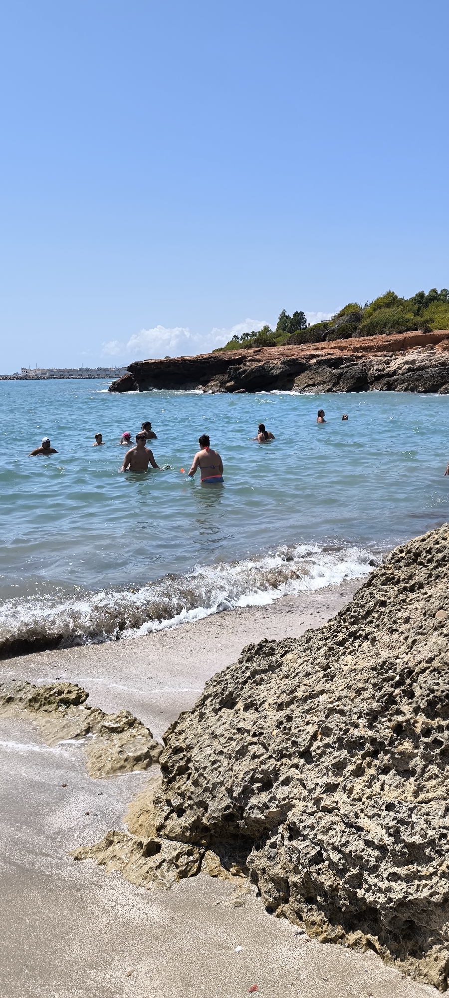 The image shows a bright, sunny day at a coastal area in Alcalà de Xivert, Spain. Several people are swimming and wading in the clear, blue ocean water. In the foreground, there are large, textured rocks and a sandy beach with small waves lapping at the shore. In the background, a rocky cliff face covered in vegetation slopes down to the sea, with a few scattered clouds in the bright blue sky. The water is calm and inviting, and the scene appears to be a popular spot for recreation. The overall impression is one of a pleasant summer day at the beach.
