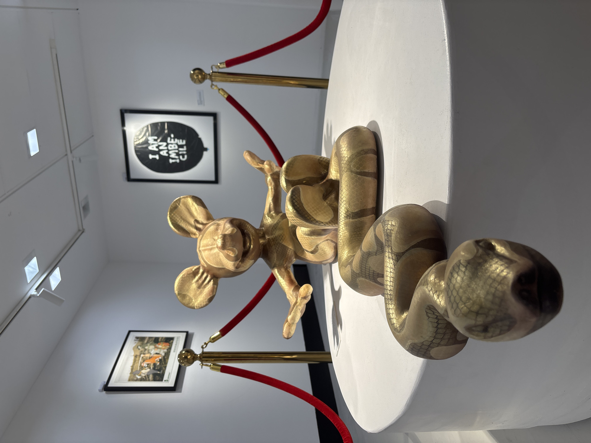 The image shows a golden sculpture of a Mickey Mouse figure intertwined with a snake, displayed on a white cylindrical platform. The sculpture is cordoned off by gold stanchions with red velvet ropes. The background features artwork on the wall, including a framed piece with the text "I AM AN IMBECILE" and another framed picture below it. The scene appears to be inside an art gallery with white walls and recessed lighting. The photograph was taken in London, United Kingdom.