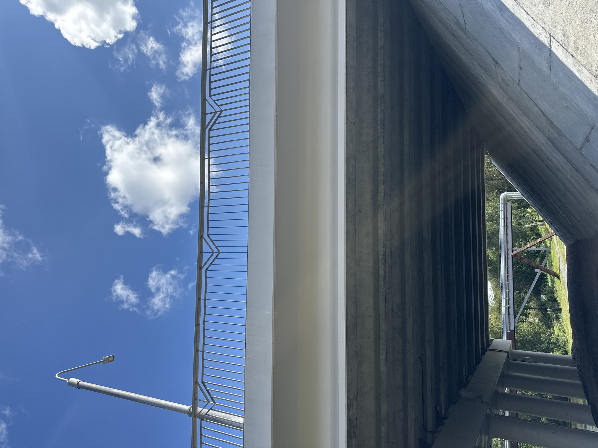 This is an outdoor, ground-level, upward-facing, wide-angle shot of a bridge under a bright blue sky with scattered white clouds. A metal railing with vertical bars and a decorative zigzag pattern runs along the top of the bridge. A modern, curved street lamp is visible on the left side, extending over the railing. The underside of the bridge is made of concrete beams, with a large, sloped concrete embankment on the right. Through the opening between the bridge supports, another smaller bridge or overpass with pipes running along it can be seen in the distance, surrounded by green trees. The scene appears to be in a park or natural area within Minsk, Belarus, as indicated by the presence of the infrastructure and the surrounding greenery. There are no people or animals visible in the image.