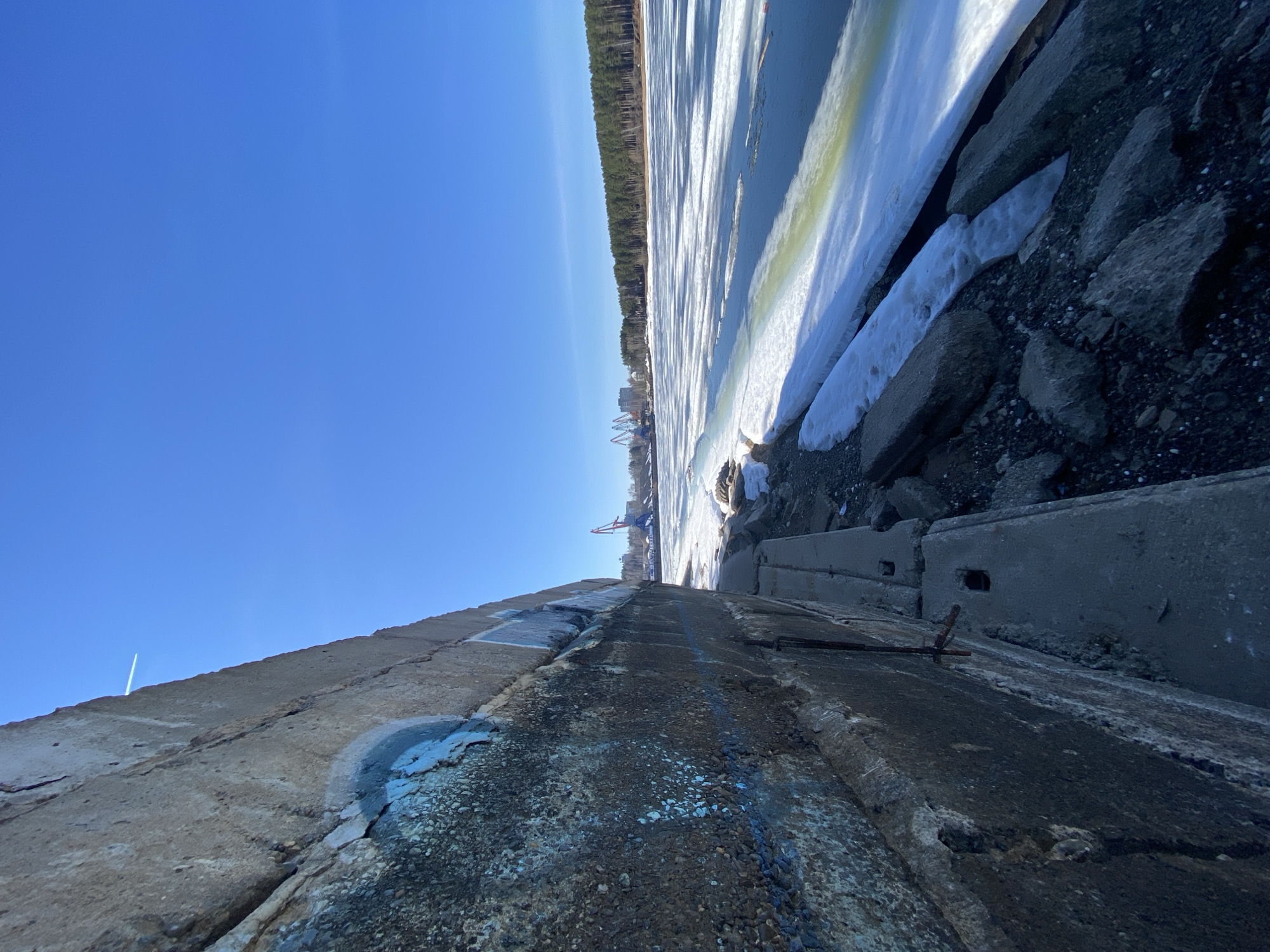 The image captures a view from a concrete embankment, likely a breakwater or pier, in the city of Chaikovsky, Russia. The perspective is angled, looking out towards a frozen body of water, possibly the Kama River, where ice and snow are visible. On the opposite shore, a line of evergreen trees is present, defining the horizon. Industrial cranes are visible in the distance near the shoreline, suggesting a port or industrial area. The sky above is clear and blue. The concrete structure in the foreground shows signs of aging, with visible cracks, discoloration, and some graffiti or paint marks. Debris and rock fragments are scattered near the edge of the embankment where it meets the ice.