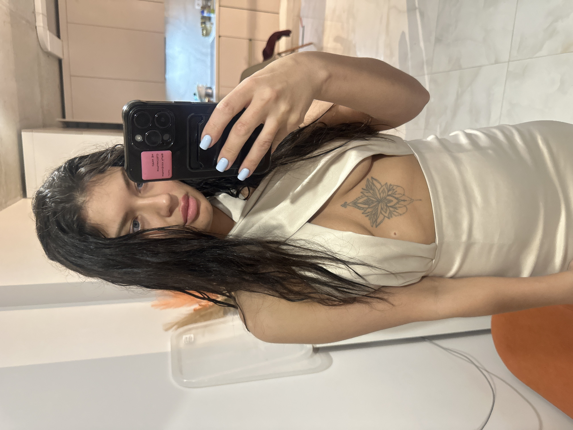 A mirror selfie captures a young woman in an indoor setting, likely a home, possibly a kitchen or bathroom, given the reflective surfaces and white cabinetry visible in the background. She holds a black smartphone with multiple camera lenses in her right hand, partially obscuring her face, and looks directly at her reflection.
The woman has long, dark, wavy hair cascading over her shoulders and chest. Her eyes are blue, and her lips have a natural pink hue. She is wearing a light-colored, possibly white or cream, satin or silky top with a V-neckline, revealing a detailed lotus-like tattoo on her sternum. Her fingernails are painted with light blue polish.
A pink sticker or card is affixed to the back of her phone case. Due to the mirror image, the text on it appears reversed and is difficult to fully decipher, but it seems to consist of capitalized letters, potentially non-English or a stylized word.
The immediate foreground and background show white countertops or surfaces. To her left, a clear plastic container is visible on the counter. To her right, a portion of an orange object, possibly a chair or cushion, is partially in view. The lighting suggests it is daytime.