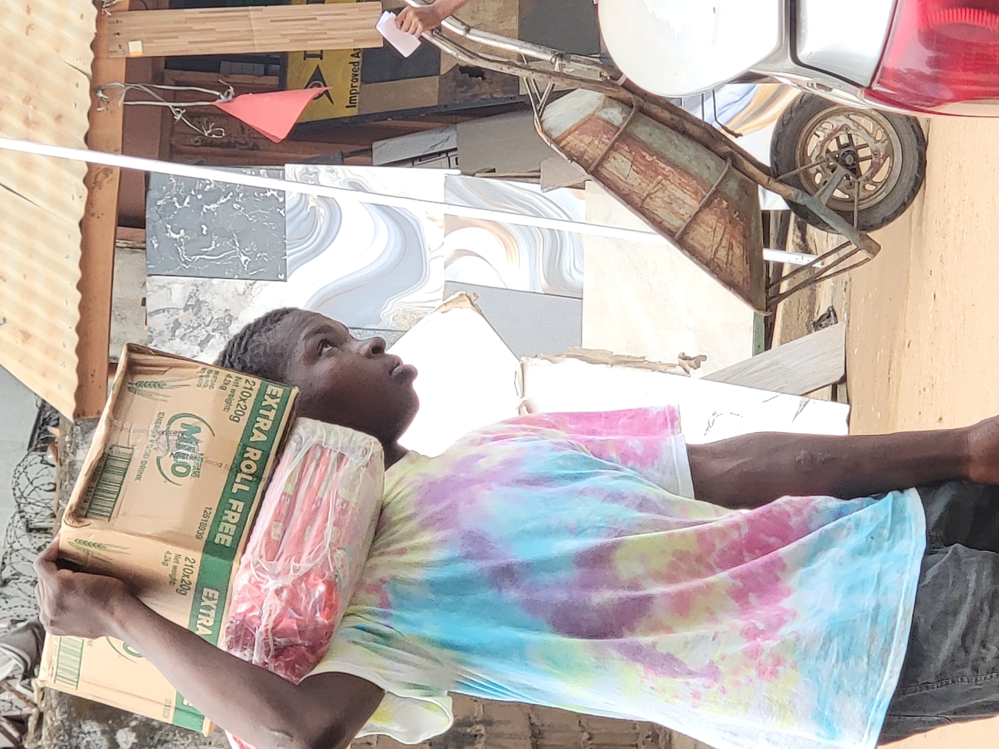 A young person with dark skin stands under a complex assortment of building materials and a vehicle part, carrying a cardboard box of Milo and a bag of items on their head. They are wearing a tie-dye t-shirt. In the background, there are tile samples, wooden planks, and a rusted wheelbarrow-like structure attached to the rear of a vehicle. The Milo box, with clear text indicating "EXTRA ROLL FREE" and "ENERGY FOOD DRINK" suggests the location is Nigeria. While Maiburiji, Nigeria is mentioned, the exact location is indeterminate from the provided image.