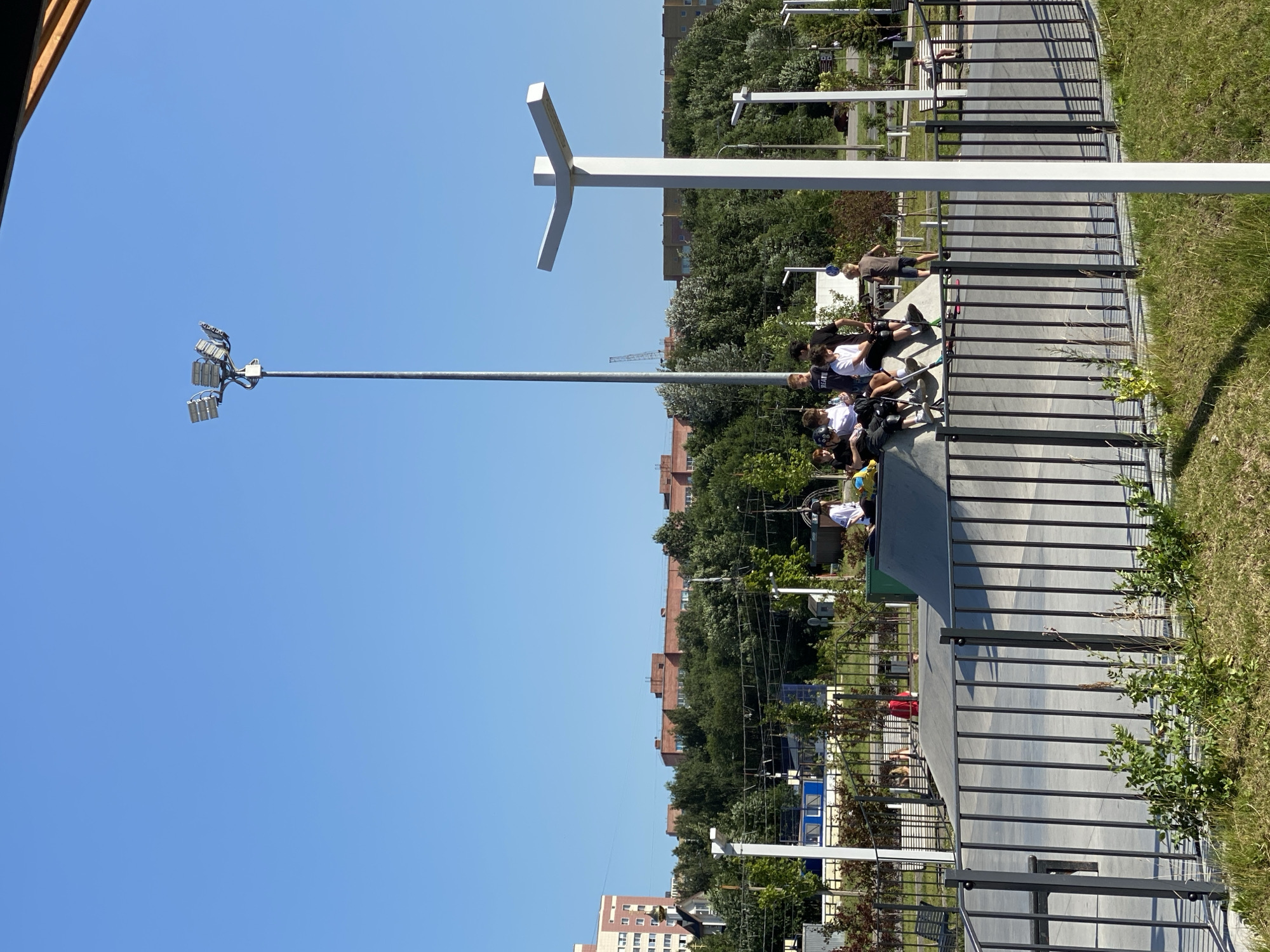 This media file depicts an outdoor scene in Cherepovets, Russia, likely on a sunny day. The main subjects are a group of young people, possibly teenagers, gathered in what appears to be a skate park or an urban recreation area. Some are seated on the edge of a ramp structure, while others are standing or preparing to use scooters or skateboards, suggesting an activity like skateboarding or scootering.
The setting includes a concrete skate park with various ramps and ledges, bordered by metal railings. Beyond the skate park, there are trees, grassy areas, and residential buildings visible in the background, consistent with an urban environment. A tall lamp post with multiple floodlights is prominent in the left portion of the frame, indicating the area might be lit for evening use. A distinctive white modern sculpture, resembling an abstract cross or an arrow, stands against the clear blue sky, adding an artistic element to the location. The presence of power lines and a distant construction crane hints at ongoing development or infrastructure in the vicinity.
The weather appears to be fair and bright, with a clear blue sky dominating the upper portion of the image, suggesting it is daytime. The clothing of the individuals, mostly t-shirts and shorts, further supports this. There is no readily discernible text visible in the image. The overall mood seems relaxed and youthful, with people enjoying outdoor activities.