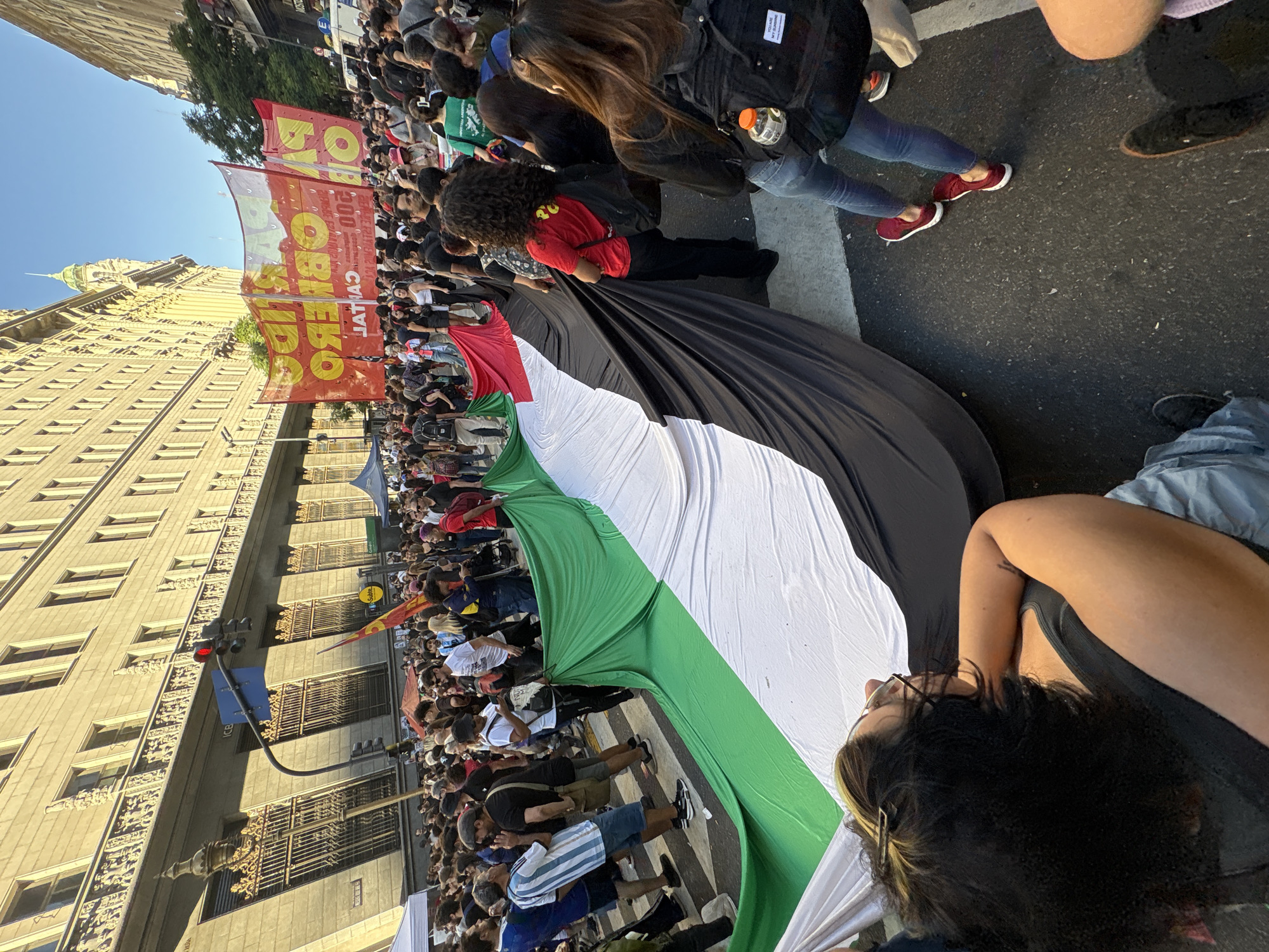 A mass demonstration is depicted on Avenida de Mayo in Buenos Aires, Argentina, with the perspective generally facing west toward the National Congress building, whose green dome is visible in the distance. The asphalt street is filled with several hundred individuals, flanked by tall, multi-story historic buildings. The building on the left features ornate balconies, arched windows with decorative metal grates, and the letters "ICE" visible on its facade. A red traffic light hangs on the left.
At the center and foreground, numerous participants are actively holding and unfolding a very large Palestinian flag, its horizontal green, white, black, and red sections extending across the width of the street. Individuals are positioned along its length, manipulating the fabric. In the background, above the crowd, two large red and yellow banners are displayed, featuring Spanish text, including "POR OBRERO" and "CATAL," indicating a political or labor-related protest. Small white and red tents are present on the left side of the street. The scene is illuminated by daylight under a clear sky, depicting an active urban protest.
