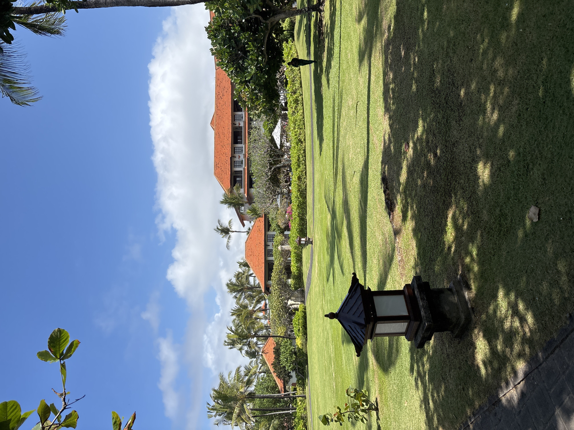 The image captures a lush green lawn in front of a resort complex in Kuta Selatan, Indonesia. The resort features multiple buildings with red tiled roofs, surrounded by palm trees and other tropical vegetation. In the foreground, a traditional stone lantern stands on the lawn, casting a shadow. A person is walking in the mid-ground towards the right side of the image, and scattered white umbrellas are visible in the distance, suggesting an outdoor seating area. The sky is a vibrant blue with scattered white clouds, indicative of fair weather. The overall scene conveys a tranquil and well-maintained resort environment.