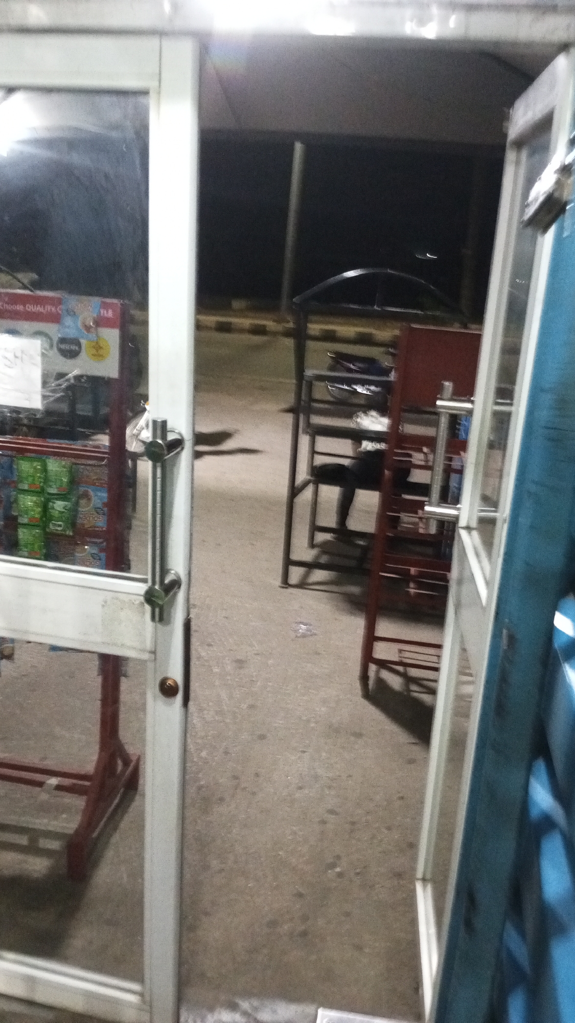 The image is captured from inside a building, looking outwards through a glass door and frame. On the left, inside the building, a display shelf is visible through the glass, laden with various packaged goods, possibly snacks or beverages. Visible text on packaging includes "CHOICE QUALITY C", "NESCAFE" logo, and "SH", along with partial text "TLE".
The primary glass door features a white frame and a prominent metallic pull handle. To the immediate right, another vertical structure, appearing to be part of a door or frame, is visible with a blue section and a white frame alongside it.
Beyond the doorway, the scene opens to an outdoor concrete or asphalt ground. In the middle ground, there are several dark metal racks or shelves, and a reddish-brown rack. A dark object resembling the seat and handlebars of a motorcycle or scooter rests on one of the dark metal racks. In the distant background, a yellow and black striped curb is discernible, suggesting a road or parking area.
The overall scene indicates it is nighttime or very late evening, as the areas outside the brightly lit immediate vicinity are dark, relying on distant artificial light sources. No specific activity is taking place, presenting a still view of a shop entrance. The setting, including the curb and shop type, is consistent with a commercial area in Adankolo, Nigeria. No emotions are discernible.