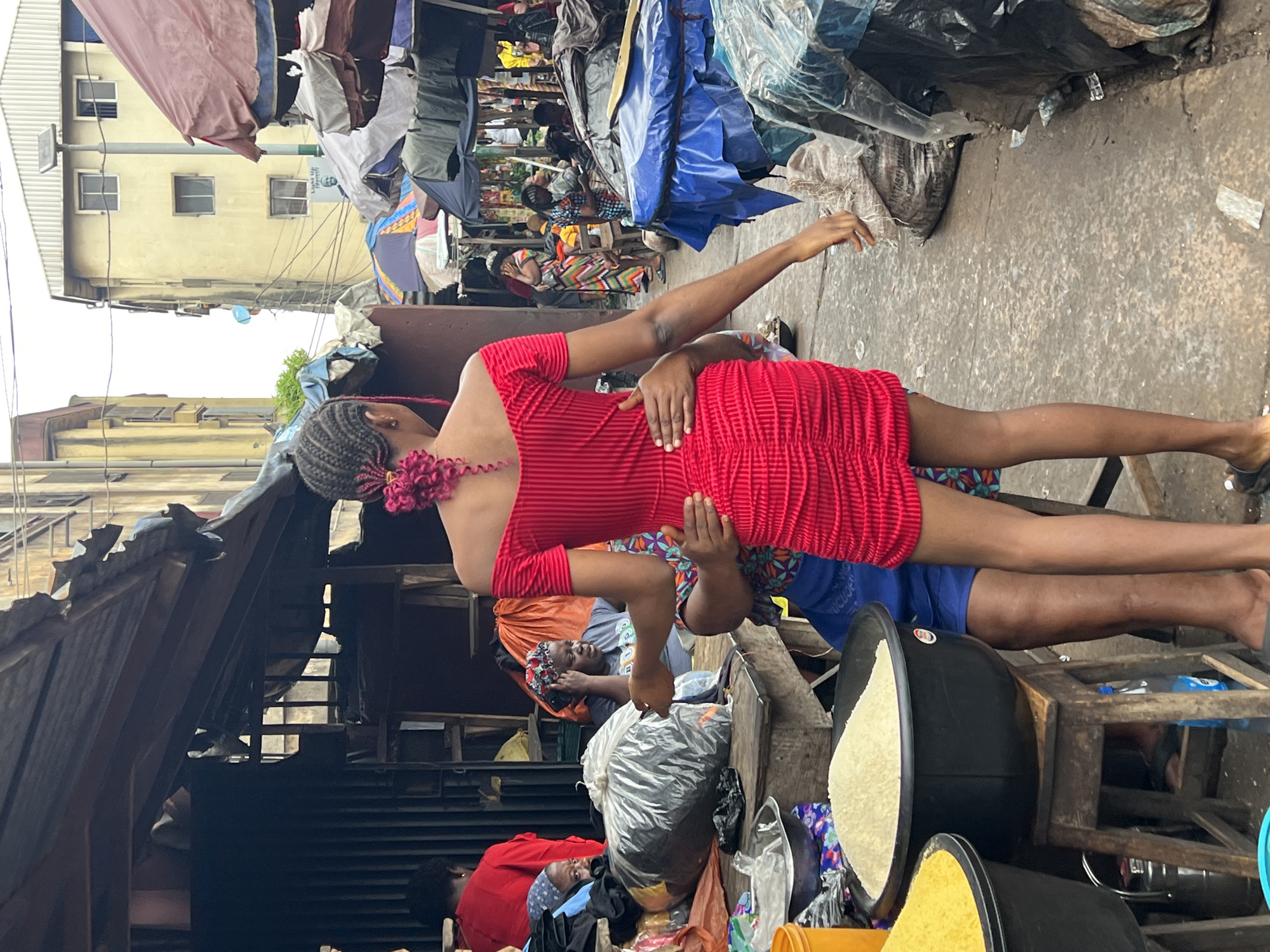 In a bustling street market in Nkpor, Nigeria, a young woman in a vibrant red, off-the-shoulder dress stands with her back to the camera, her dark braided hair adorned with pink ties. She is being embraced from behind by another person whose hands are placed on her waist, one of which is visible. The woman in red's right hand is extended slightly forward. The scene unfolds in an open-air market with stalls and merchandise lining a crowded walkway. In the foreground, large black bowls filled with white and yellow grains, likely rice, are displayed on wooden stools. Behind the central figures, market activities are in full swing. Stalls are covered with tarps and fabrics, displaying a variety of goods including bags of produce, textiles, and other assorted items. Buildings with multiple windows, some painted yellow and others appearing older and weathered, form the backdrop of the market. People are seen moving through the market, interacting with vendors, and browsing merchandise. The overall atmosphere suggests a typical day in a busy Nigerian marketplace.