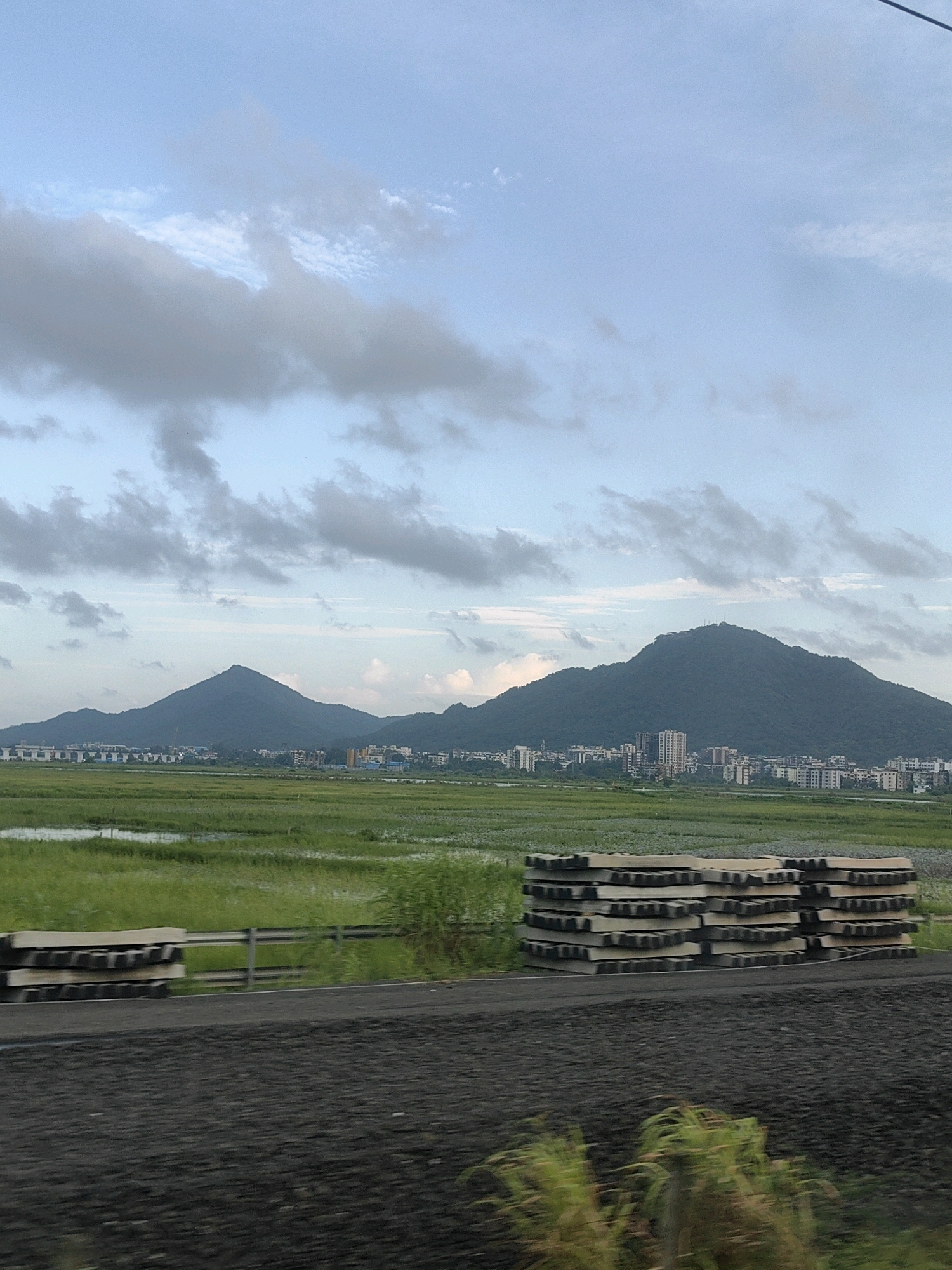 The image shows a landscape scene under a cloudy sky, featuring a town nestled at the foot of a verdant mountain range. In the foreground, a stack of what appear to be railway sleepers or concrete beams are arranged. Beyond this, a vast expanse of green fields stretches out, interspersed with areas of water. The town itself consists of numerous buildings, including apartments and other structures, built close together. The mountain is covered in dense vegetation, and a peak with some structures is visible at its summit. The overall impression is one of a rural or semi-rural setting with developing infrastructure.