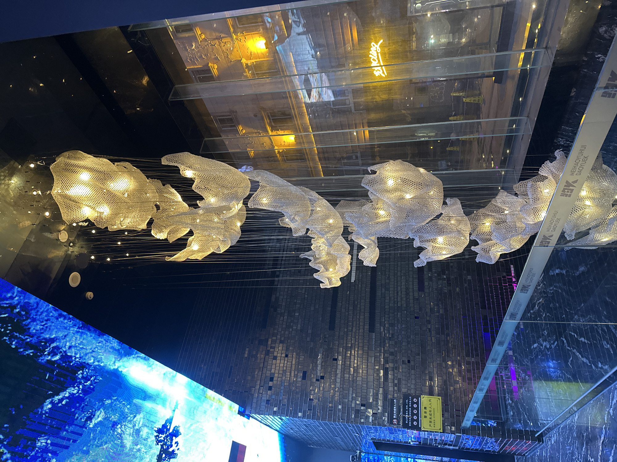 Here is a description of the image:
The photograph, taken at eye level, presents an interior view of what appears to be a modern, possibly commercial, space in Liangjiang, China. The scene is dominated by an elaborate, illuminated chandelier or art installation composed of interconnected, cloud-like or floral forms made from a perforated material that glows with internal lights.
The ceiling above features a glossy, reflective surface, possibly made of glass or polished material, mirroring the elements below and creating an illusion of depth. Reflected in the ceiling is an exterior scene, possibly a building facade at night, illuminated with lights, adding a layer of complexity to the composition.
To the bottom left of the frame, a portion of a large digital display or screen is visible, showcasing a vibrant, abstract image of swirling blues, possibly depicting a geographical landscape or abstract art.
Along the right edge of the frame, a vertical strip with the inscription "MEIHAOYUHUI DAOYUEK" is visible, possibly indicating the location or brand. The opposite side features a warning sign written in Chinese.
The general environment appears to be well-lit and possibly located in a shopping mall or exhibition center. The composition suggests a sophisticated, contemporary design aesthetic.
