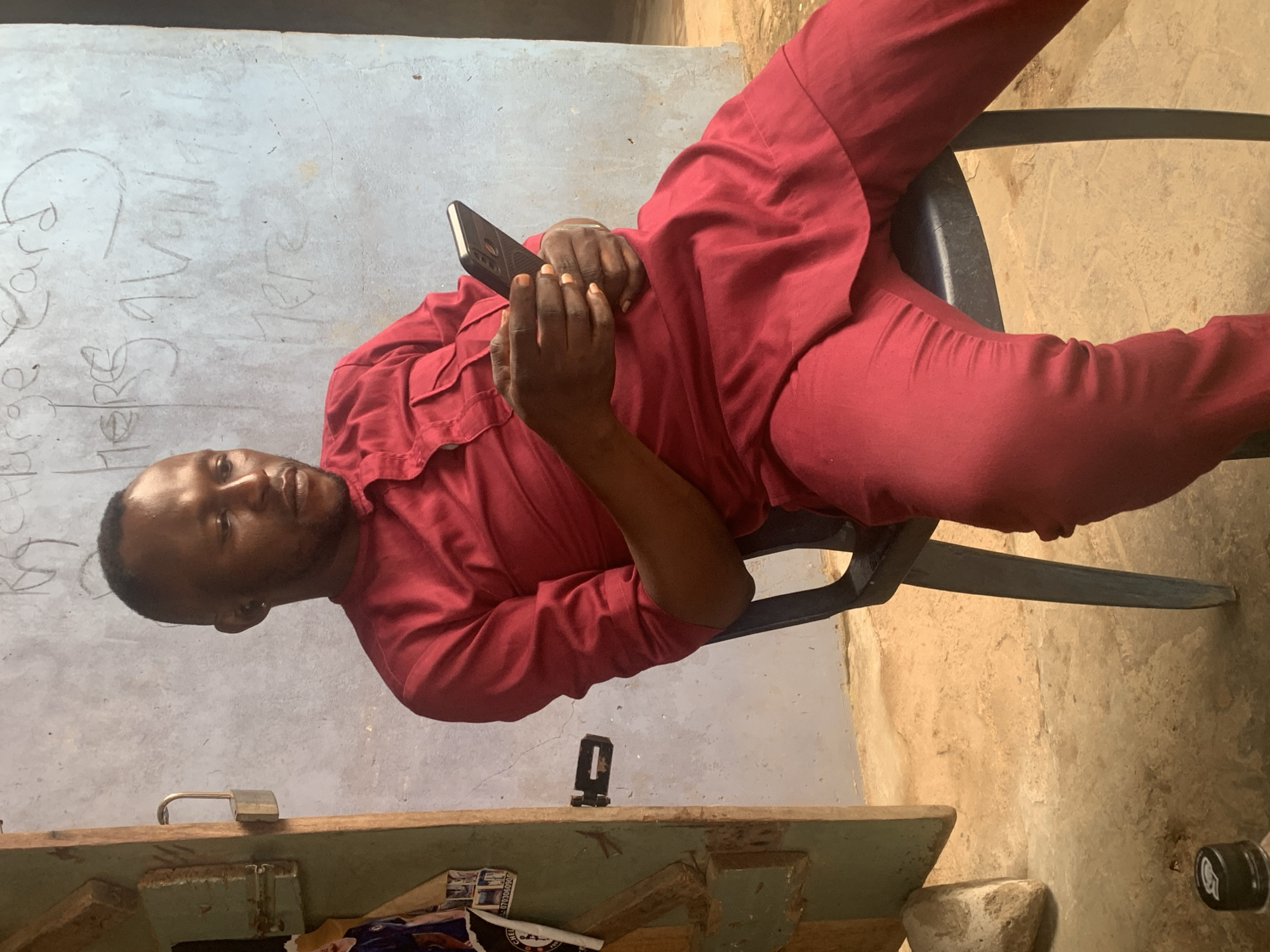 A Black man wearing a red kaftan and pants sits on a dark chair, holding a black smartphone in his hands. He is looking down at the phone. The background consists of a light blue wall with some handwritten text, and to the left, a wooden door with a padlock. The floor is concrete. The scene appears to be outdoors, possibly in a residential area or shopfront in Shagamu, Nigeria. The lighting suggests it is daytime.
