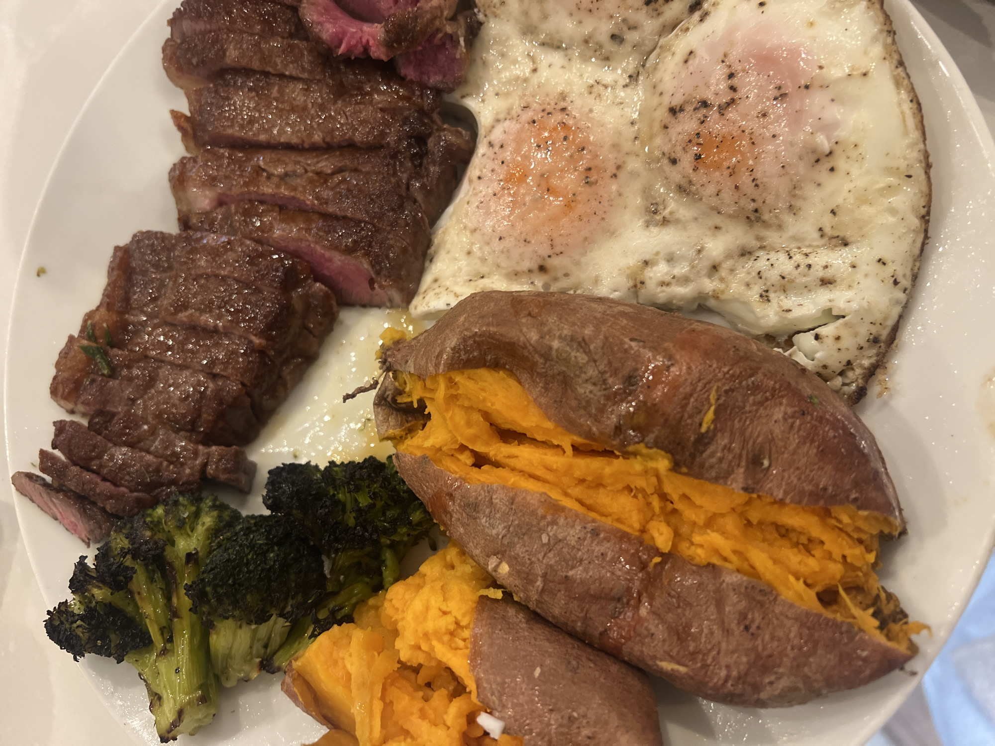 A close-up overhead shot displays a hearty meal served on a white plate. The main components are sliced steak, two fried eggs, sweet potatoes, and broccoli.
The steak, sliced against the grain, shows a pink, medium-rare interior, suggesting it's tender and juicy. Some slices have a light char on the exterior. Adjacent to the steak are two fried eggs, seasoned with black pepper, their yolks slightly visible through the cooked whites, indicating they are likely sunny-side up or over easy.
In the lower half of the plate, two sweet potatoes are prominently featured. They appear baked or roasted, split open to reveal their vibrant orange, soft flesh. One of the sweet potatoes is partially mashed within its skin. A serving of steamed or roasted broccoli florets, some with slightly charred edges, accompanies the sweet potatoes, adding a green vegetable component to the meal.
The presentation suggests a wholesome, protein-rich dish. The lighting appears to be indoor artificial light. No people or text are visible in the image.