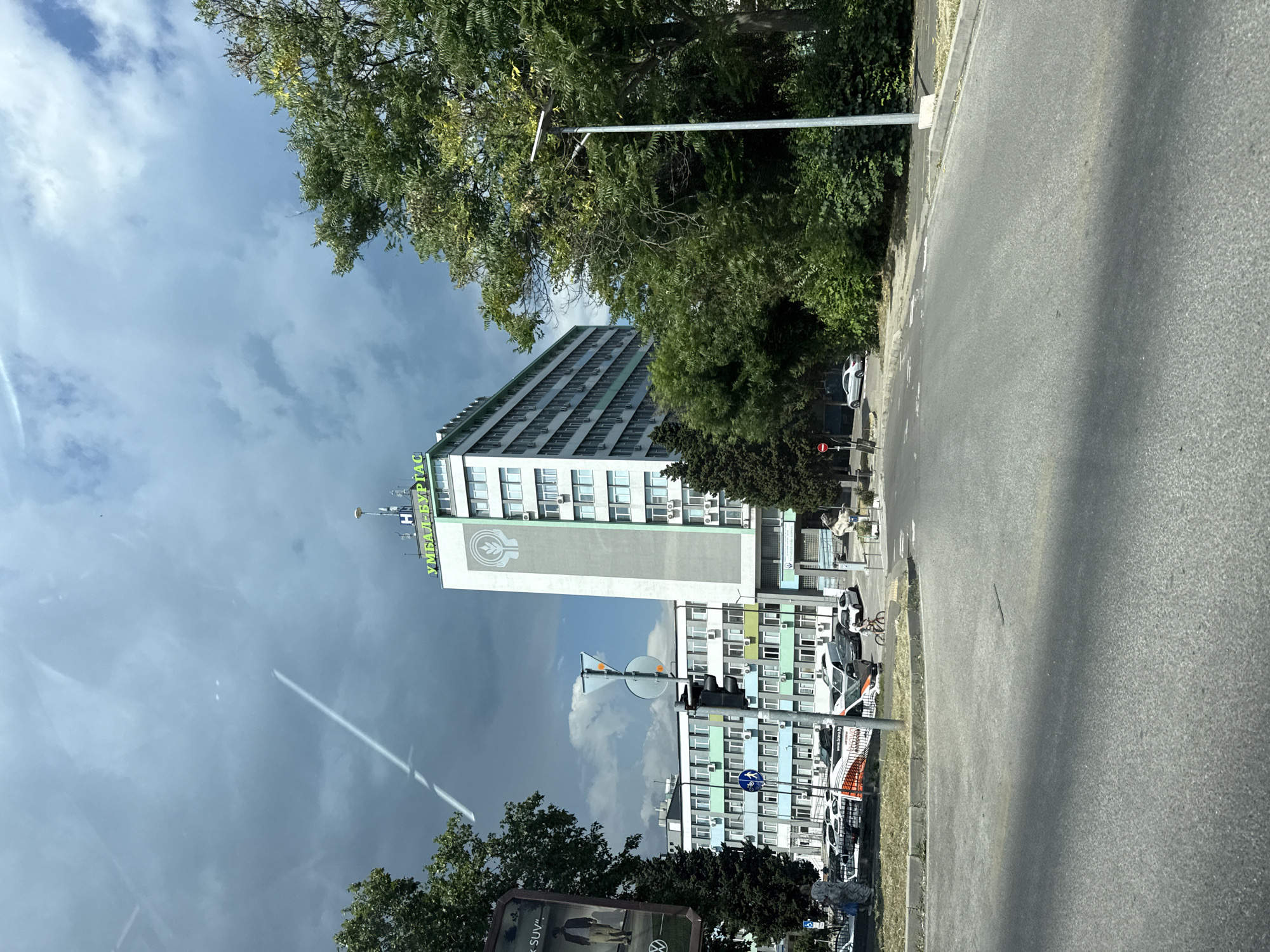 The image depicts a street scene in Burgas, Bulgaria, with a prominent multi-story building in the background. The building, identified by green lettering at the top as "УМБАЛ-БУРГАС" (UMBAL-BURGASS, meaning University Hospital Burgas), has a light-colored facade with numerous windows and air conditioning units. A large circular emblem is visible on the side of the building. To the left of this main building, another multi-story structure with a light blue and green facade is present, also featuring many windows. The foreground shows a street with a slight incline, a paved road, and a concrete curb. Several cars are parked along the street and in a lot in front of the buildings. A person on a bicycle is visible in the mid-ground, riding on the road. Trees with green foliage line the street and surround the buildings, and a street lamp stands on the right side of the image. The sky above is partly cloudy, with some dramatic dark clouds and a distinct white contrail from an aircraft. A traffic light is visible on a pole in the mid-ground, and various street signs are present, including pedestrian and traffic directional signs. A billboard advertisement is partially visible on the far left of the image.