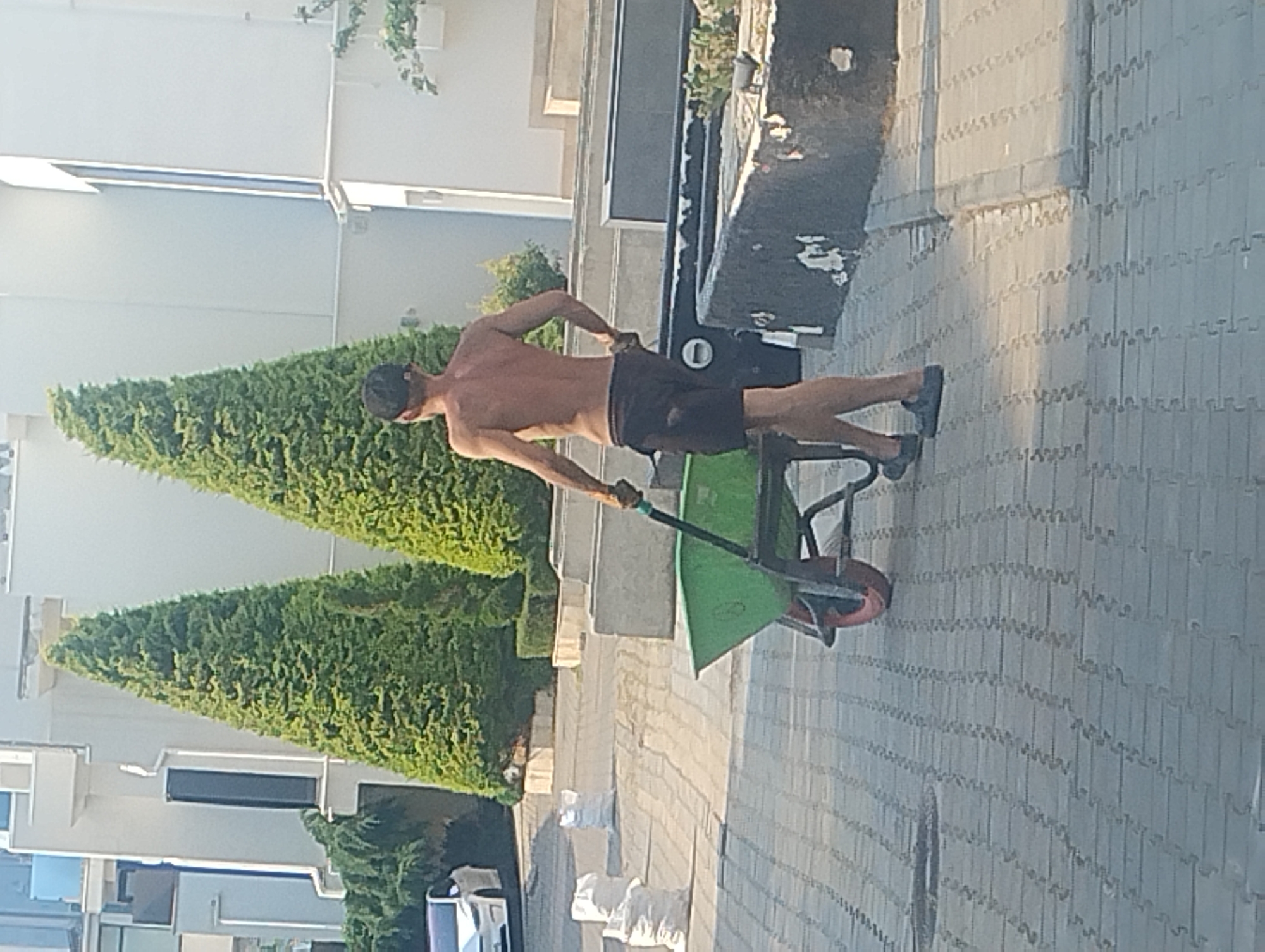 A shirtless man wearing black shorts and a dark cap is pushing a green wheelbarrow on a paved walkway. He is outdoors, with manicured evergreen trees on his left and a modern building on his right. The sun is shining brightly, casting shadows and indicating it is likely daytime. The man appears to be engaged in some kind of work, possibly gardening or landscaping, as he is using the wheelbarrow. The paved walkway has a pattern of interlocking grey stones. There are white objects, possibly bags, scattered on the walkway behind the wheelbarrow. The building in the background has a white facade and appears to be a multi-story structure with large windows. The overall scene suggests a warm, sunny day in a residential or commercial area with landscaping.
