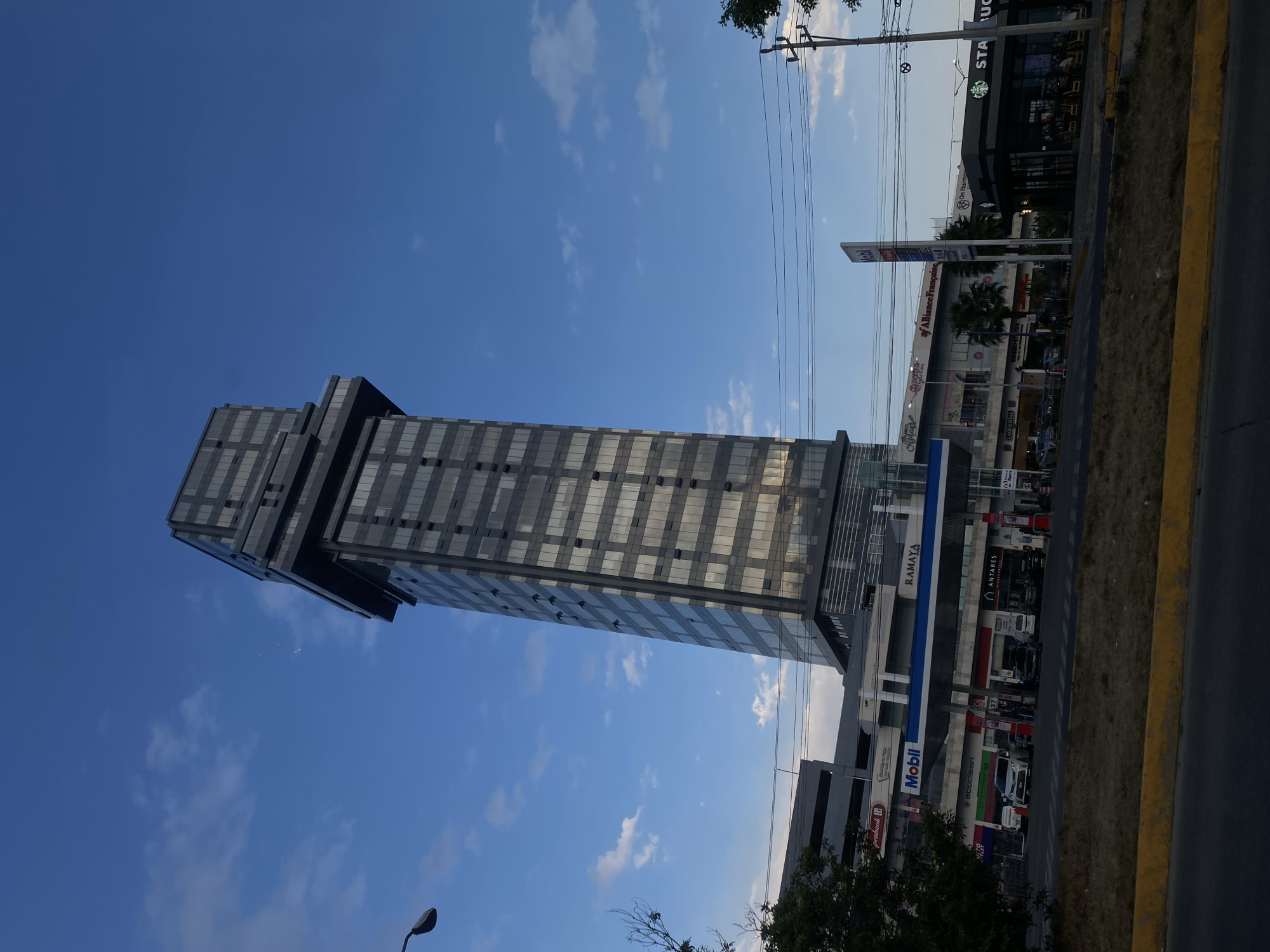 The image captures a modern building in San Bernardino Tlaxcalancingo, Mexico. The building is tall and narrow, with a gray exterior and multiple windows. The sky is blue with scattered clouds. A Starbucks is located to the right of the building, along with a Mobil gas station. The angle of the photo makes the building appear to be leaning to the left. There is a sidewalk and a street adjacent to the building.