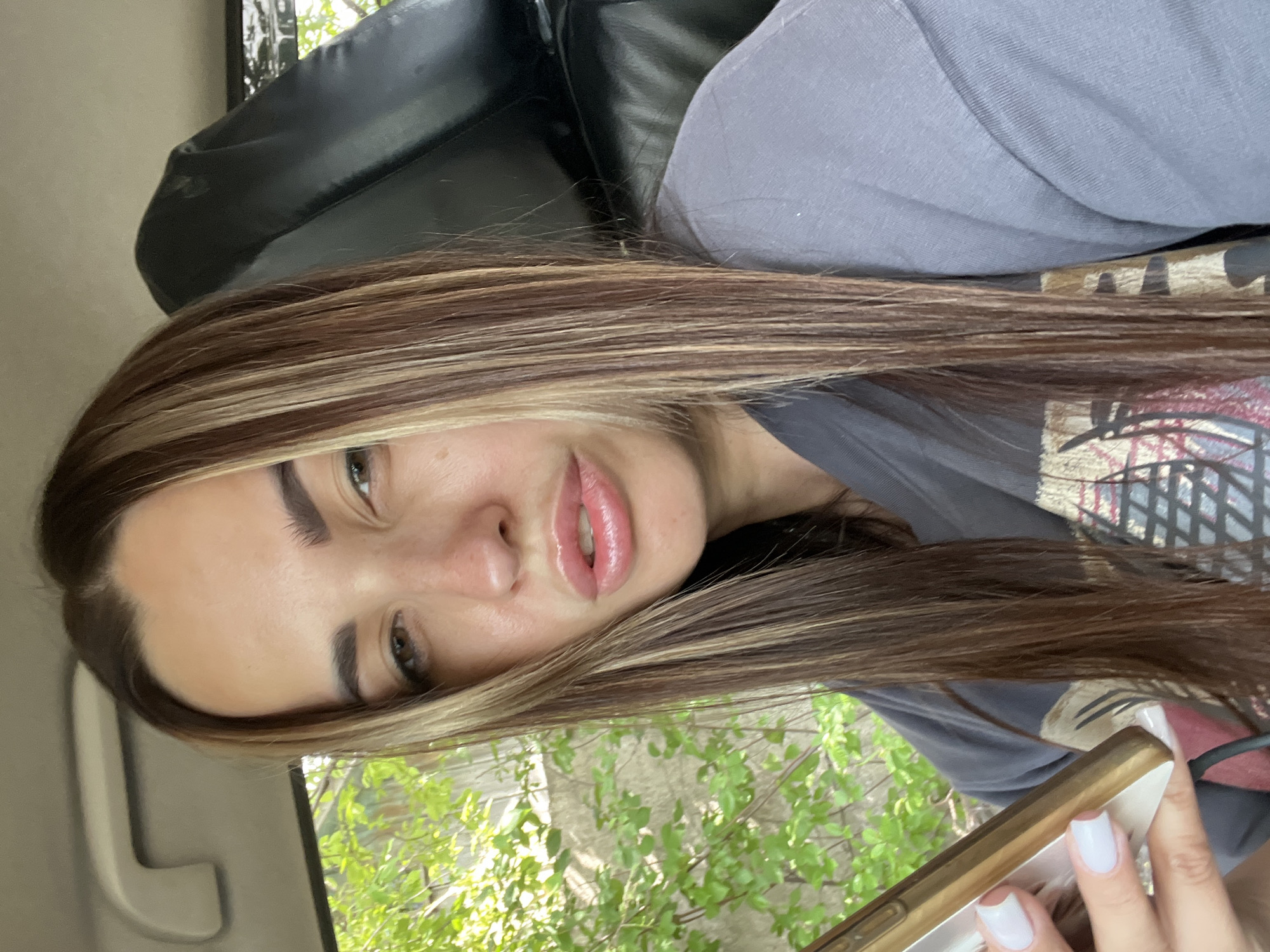 The image shows a close-up, sideways selfie of a young woman with long, brown hair with blonde highlights. She is wearing a dark t-shirt. Her face is partially obscured by her hair. She has prominent eyebrows and appears to be wearing lip gloss. She is holding a smartphone in her right hand, which has white nail polish. The background suggests she is inside a vehicle, with a black seat behind her and a glimpse of green foliage and a wooden fence through a window. The lighting suggests it is daytime.
