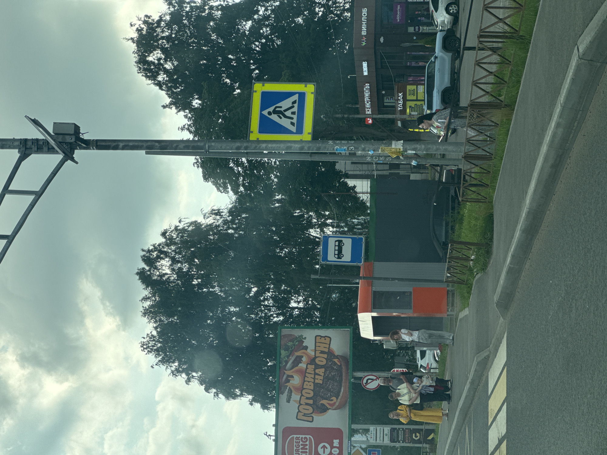 The image shows a street scene in Novoe Devyatkino, Russia. A pedestrian crossing is visible on the right side of the road, with a yellow and blue pedestrian crossing sign above it. A bus stop sign is also visible. On the left side of the street, there are several buildings with various signs, including "WILDBERRIES," "ВИНЛАБ" (Vinlab), and "ТАБАК" (Tabak). A large billboard for Burger King is also present, displaying the slogan "Готовим НА ОГНЕ" (Cooking ON FIRE).
Several people are standing on the sidewalk near the pedestrian crossing, some appear to be waiting. The weather appears to be overcast. The overall scene suggests an urban or suburban environment with commercial establishments. The orientation of the image suggests it was taken from a vehicle moving along the road.