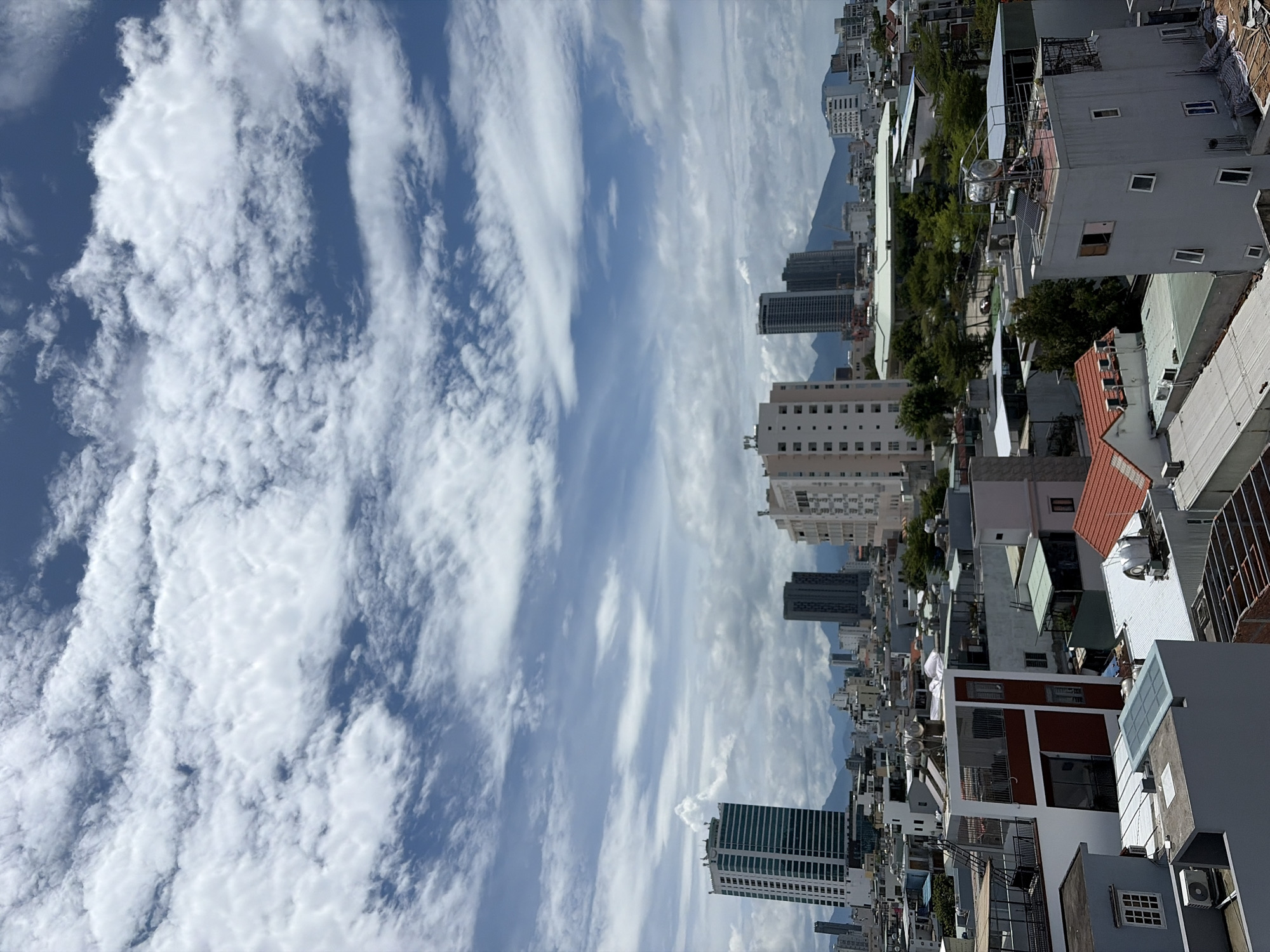 This is a high-angle shot of a city skyline in Da Nang, Vietnam, on a cloudy day. The foreground features a dense collection of residential buildings with varying architectural styles, many of which have flat roofs with visible infrastructure like water tanks, solar panels, and air conditioning units. Some buildings have red tile roofs. In the middle ground, several modern high-rise buildings are visible, including a prominent light-colored skyscraper with many windows and a darker, more slender skyscraper. Further in the background, more buildings and mountains can be seen. The sky is filled with dynamic, white cumulus clouds against a blue sky. No people are visible in the image.