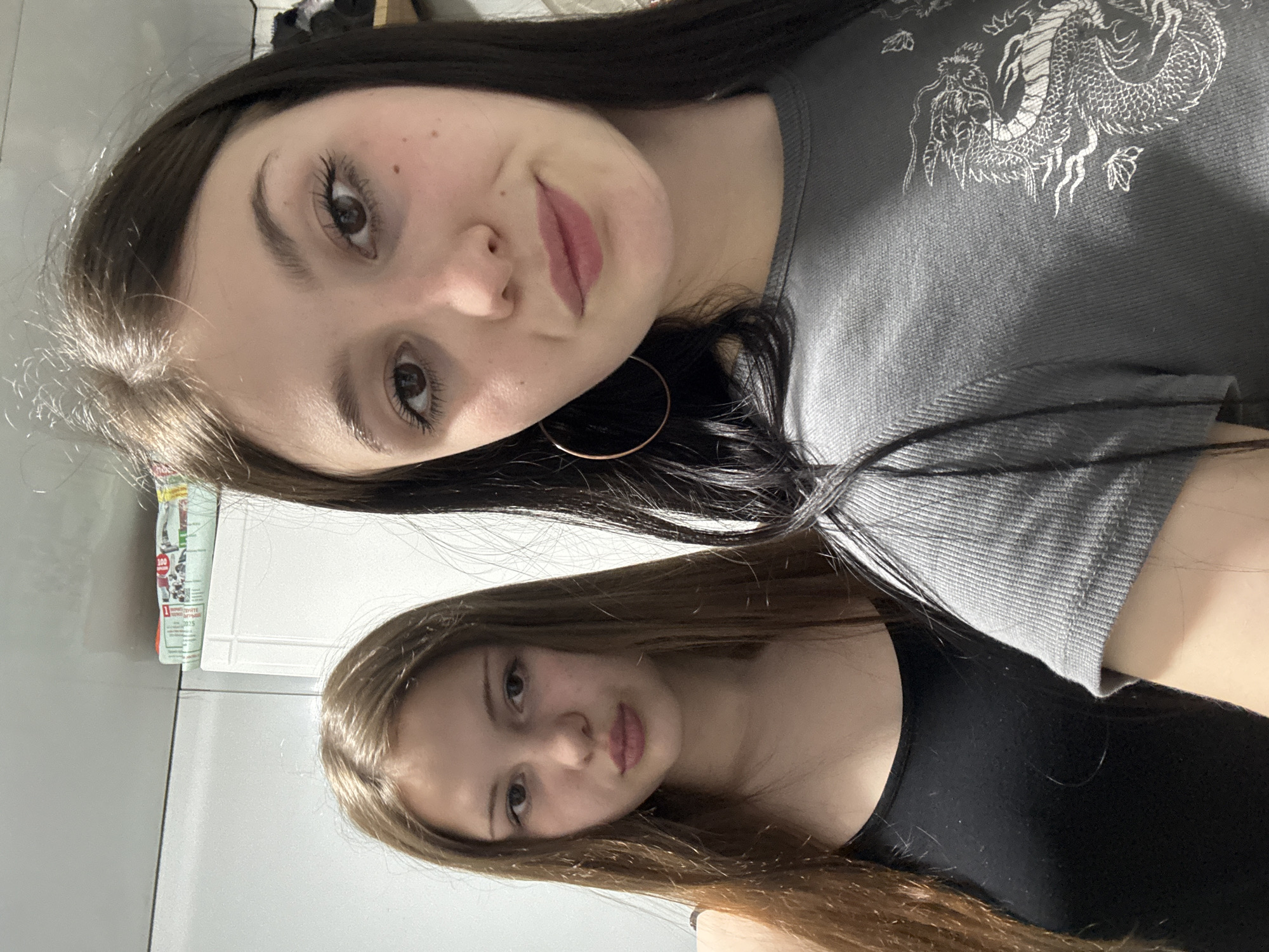 Two women are in the image. The woman on top has long dark hair and is wearing a gray shirt with a dragon design and gold hoop earrings. The woman on the bottom has long light brown hair and is wearing a black tank top. The background appears to be inside a room with white walls. A package with text in a foreign language is visible in the background, along with a white cabinet. The location is not explicitly identified.