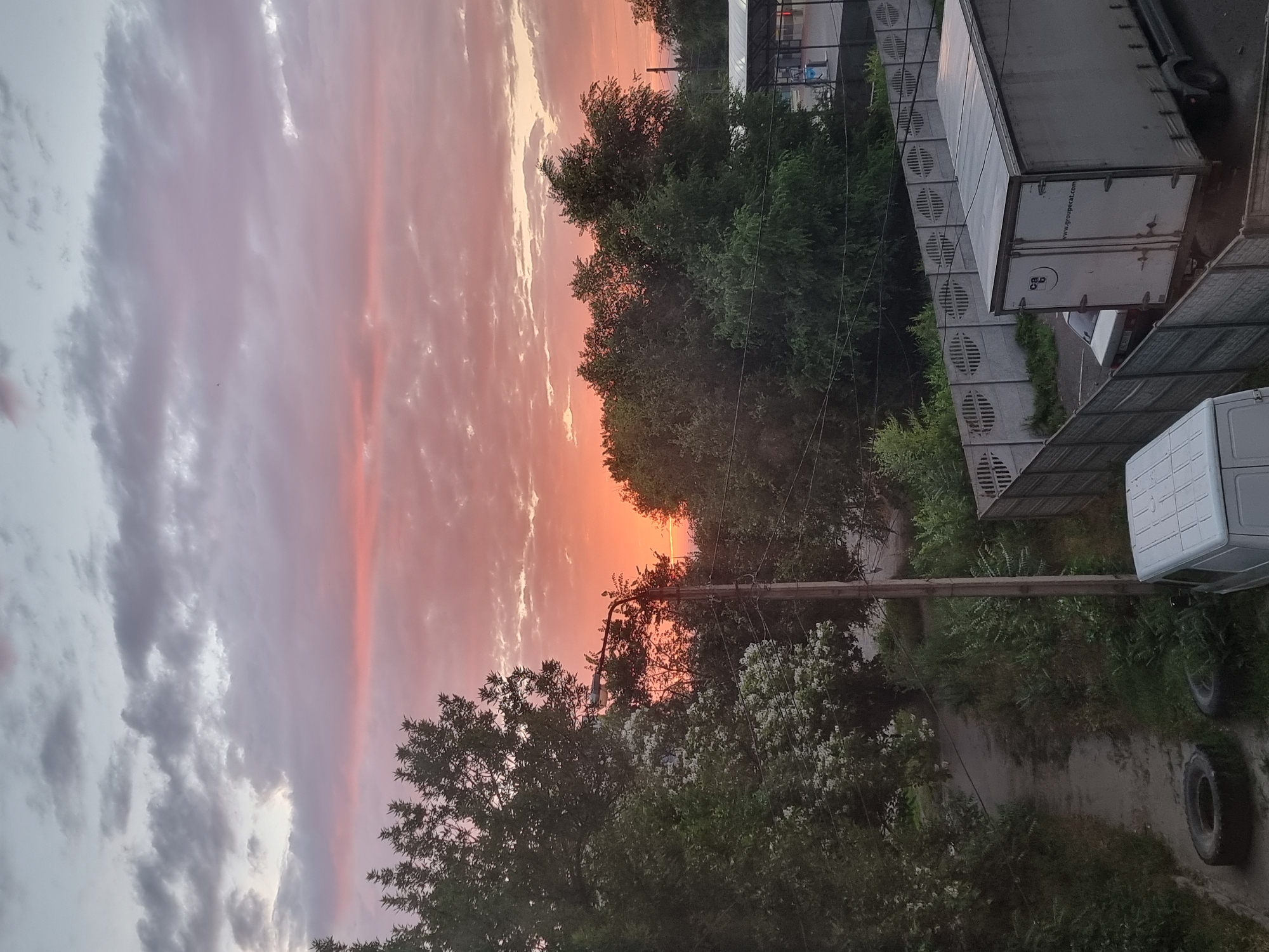 A vertical, eye-level, medium shot captures a scenic sunset over a residential area in Kryvyi Rih, Ukraine. The sky is a canvas of soft pinks and oranges, transitioning to pale blue at the top, with textured clouds scattering the light. Below the vibrant sky, a dense canopy of lush green trees fills the mid-ground, their leaves creating a dark silhouette against the colorful horizon. A single, old-fashioned street lamp stands tall on a wooden pole on the left side of the frame, its presence hinting at the urban setting. In the foreground, a narrow, unpaved path winds through overgrown vegetation. A discarded car tire lies on the path, near another tire partially obscured by grass. To the right, a grey concrete fence separates the residential area from a parking lot where a white truck with the text "www.groupecat.com" on its rear doors is parked, alongside a white van and a small car. The overall impression is one of peaceful twilight in a suburban Ukrainian landscape.