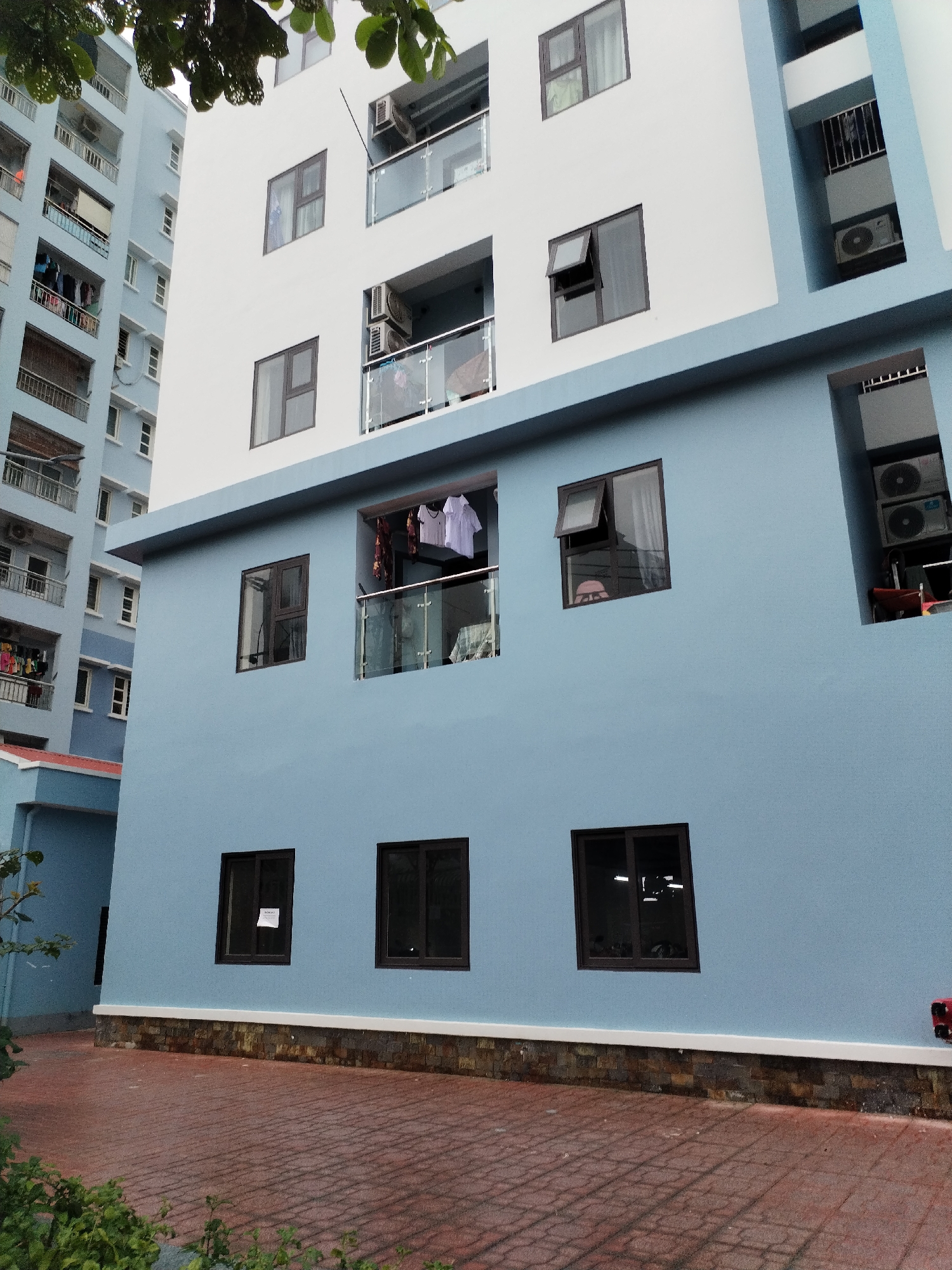 This image depicts a residential scene in Nha Trang Ward, Vietnam, featuring two multi-story apartment buildings under bright daylight.
The prominent building in the foreground is painted in two distinct sections: a light blue lower half and a white upper half. It features numerous dark-framed windows and modern glass balconies. On the blue section, an upper-level window is open, revealing white and patterned laundry hanging on a rack behind the balcony. Another open window on the same level shows the top of a person's head, possibly wearing a pink cap, partially visible inside. To the far right of this section, a recessed area houses two stacked air conditioning condenser units, with some items stored below. On the lower blue wall, one of the three visible windows has a white paper sign taped to its interior, bearing text that appears to be "NOM HUY". The base of this building is adorned with a decorative dark stone and brick border.
In the background, to the left, stands a taller, light blue apartment complex. This building also features many balconies, several of which are used for drying clothes and accommodate external air conditioning units, characteristic of urban residential life.
The ground in the foreground is covered with reddish-brown patterned paving stones, resembling a courtyard or pathway. Green foliage is visible in the bottom left corner and peeking from the top left, suggesting some surrounding greenery. The bright lighting indicates it is daytime, likely with clear or mildly overcast skies, conducive to drying laundry. The scene conveys an active, lived-in urban environment.