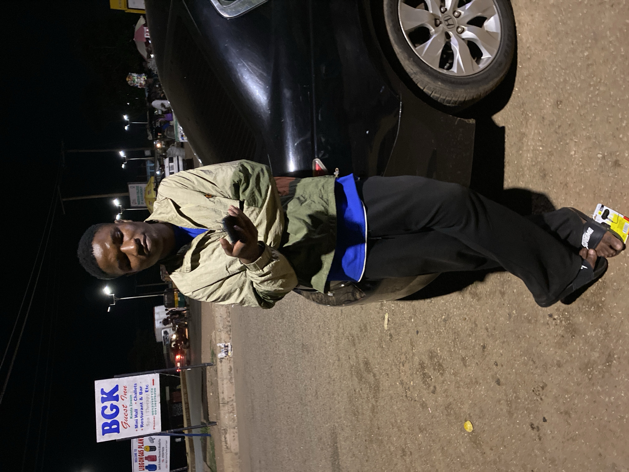 A young Black male stands at night next to a black sedan in Bukuru, Nigeria. He is wearing a tan and olive green jacket over a blue shirt and black pants, with black sandals on his feet. He is holding a black mobile phone in his right hand and looking at it. The car is parked on a dirt and asphalt road. In the background, there are streetlights, a white truck, other vehicles, people, and signs, including a prominent sign for "BGK Guest Inn." Another sign to the left reads "WELCOME TO LIGBON GAS PLANT." The overall lighting suggests it is after dark.
