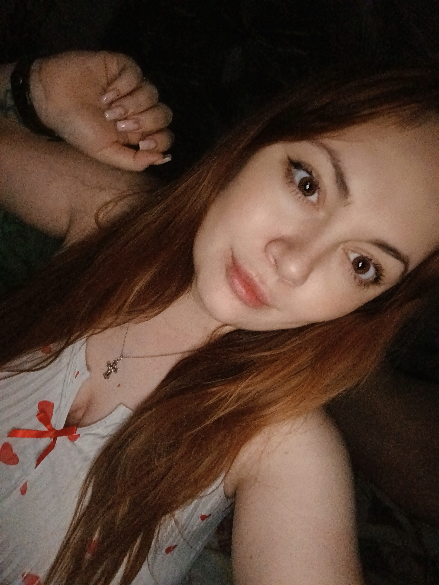 A close-up, selfie-style shot depicts a young woman with reddish-brown hair. She is wearing a light-colored top with a red bow and small heart patterns. A silver necklace with a cross pendant is visible around her neck. Her left hand, adorned with light-colored manicured nails, is raised in the upper left corner of the frame. The background is dark and indistinct. There is no verifiable information from the image to confirm the location is in Chaikovsky, Russia.