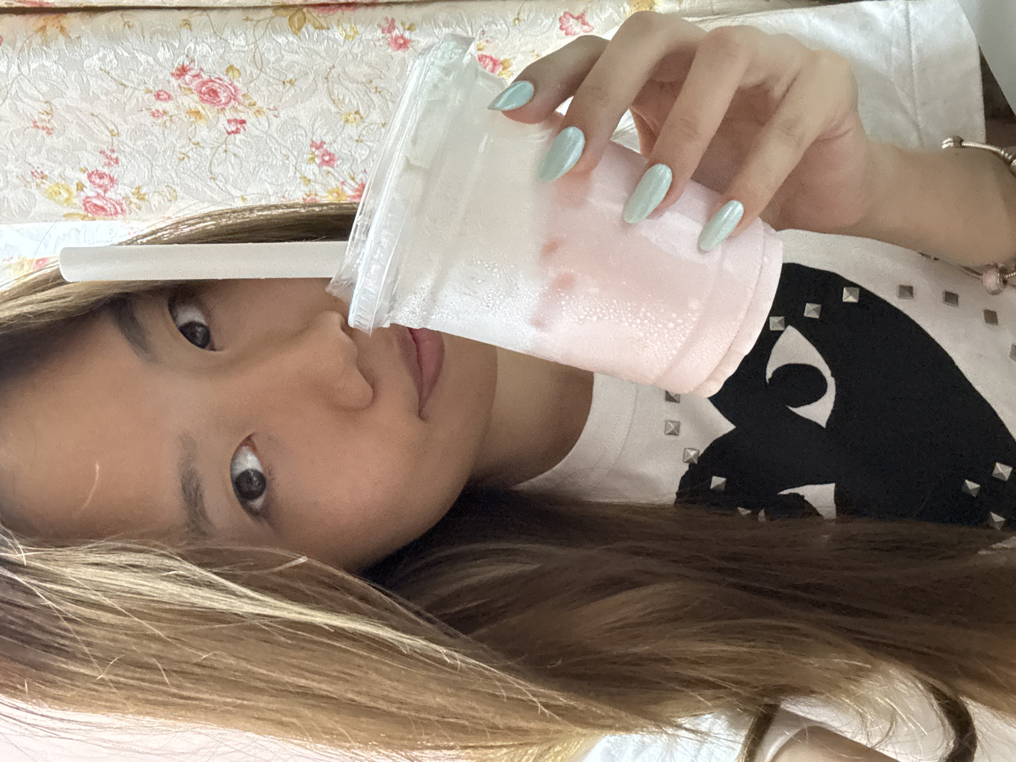 A young woman is lying on what appears to be a bed or sofa with a floral patterned cover. She is holding a large plastic cup containing a pink drink, possibly a smoothie or milk-based beverage, with a white straw. The drink has condensation on the outside of the cup. The woman has light brown hair and light blue, oval-shaped nails. She is wearing a white t-shirt with a black design that includes silver studs. The woman is looking directly at the camera. The location is in a residential space, implied to be in Shapingba District, China.