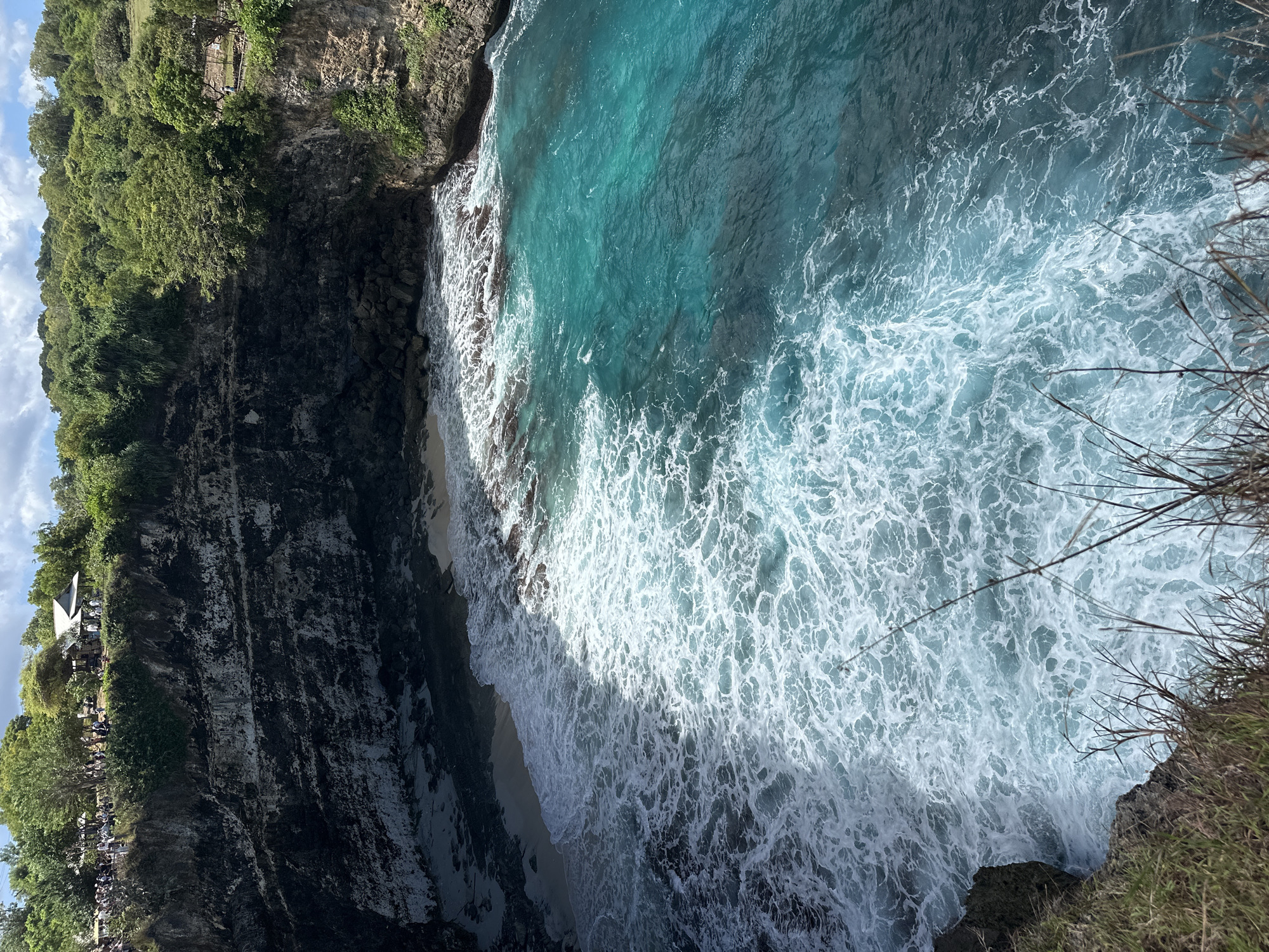 The image presents a scenic aerial view of a coastal area, likely located in Nusa Penida, Indonesia, based on the distinctive turquoise waters and dramatic cliffs. The scene is dominated by a rocky cliff face on the left side, densely covered with lush green vegetation. The cliff drops steeply towards a narrow sandy beach below, where the waves break aggressively against the shore, creating a frothy white foam that contrasts sharply with the deep blue of the ocean. The ocean surface is turbulent, with clear signs of wave action and currents. On the right side of the image, the edge of another cliff is visible, with dry grass and sparse vegetation clinging to its edge. The composition provides a high-angle, almost bird's-eye perspective of the landscape, emphasizing the scale and rugged beauty of the coastal environment. The water color varies from deep turquoise to foamy white near the shore. There is a man-made structure with white peaked roofs visible atop the cliff on the left side, suggesting the presence of a tourist lookout point. Groups of small figures are visible near the structure, indicating human presence.