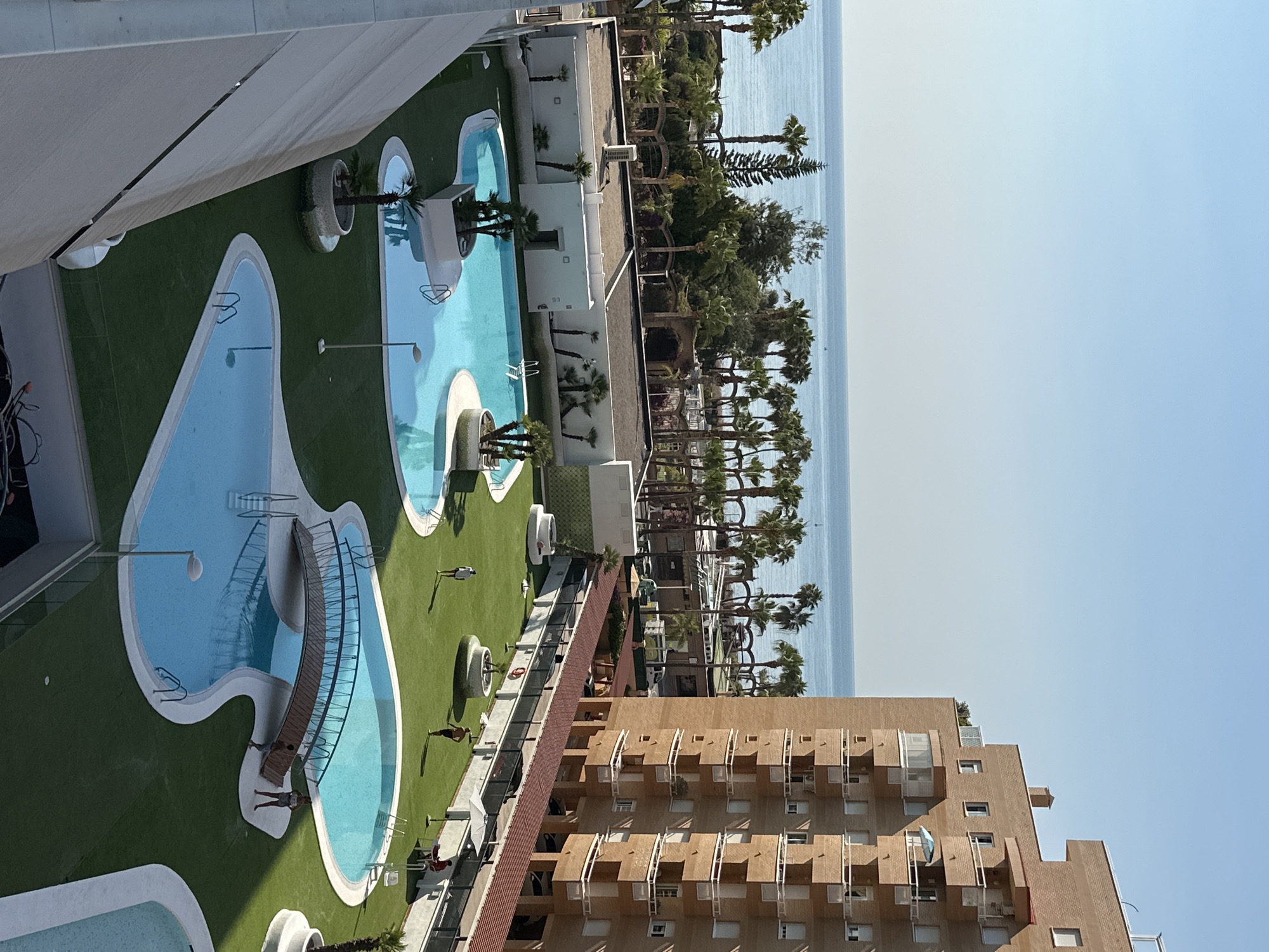 The image shows a bright, sunny day at a resort in Les Amplaries, Spain, with a view of the Mediterranean Sea. The foreground is dominated by a large, multi-level swimming pool complex with turquoise water and a decorative wooden bridge spanning one of the pools. The pools are surrounded by neatly manicured green artificial turf. Several palm trees and other tropical plants are scattered throughout the grounds. To the right, a tall, multi-story brick apartment building with numerous balconies overlooks the pool area. In the background, a line of palm trees and other vegetation separates the resort from the calm, blue sea. The sky is clear and a bright blue. Several people are visible in the image, including individuals walking on the grass, standing by the pool, and sitting at a table. One person appears to be working near the pool.