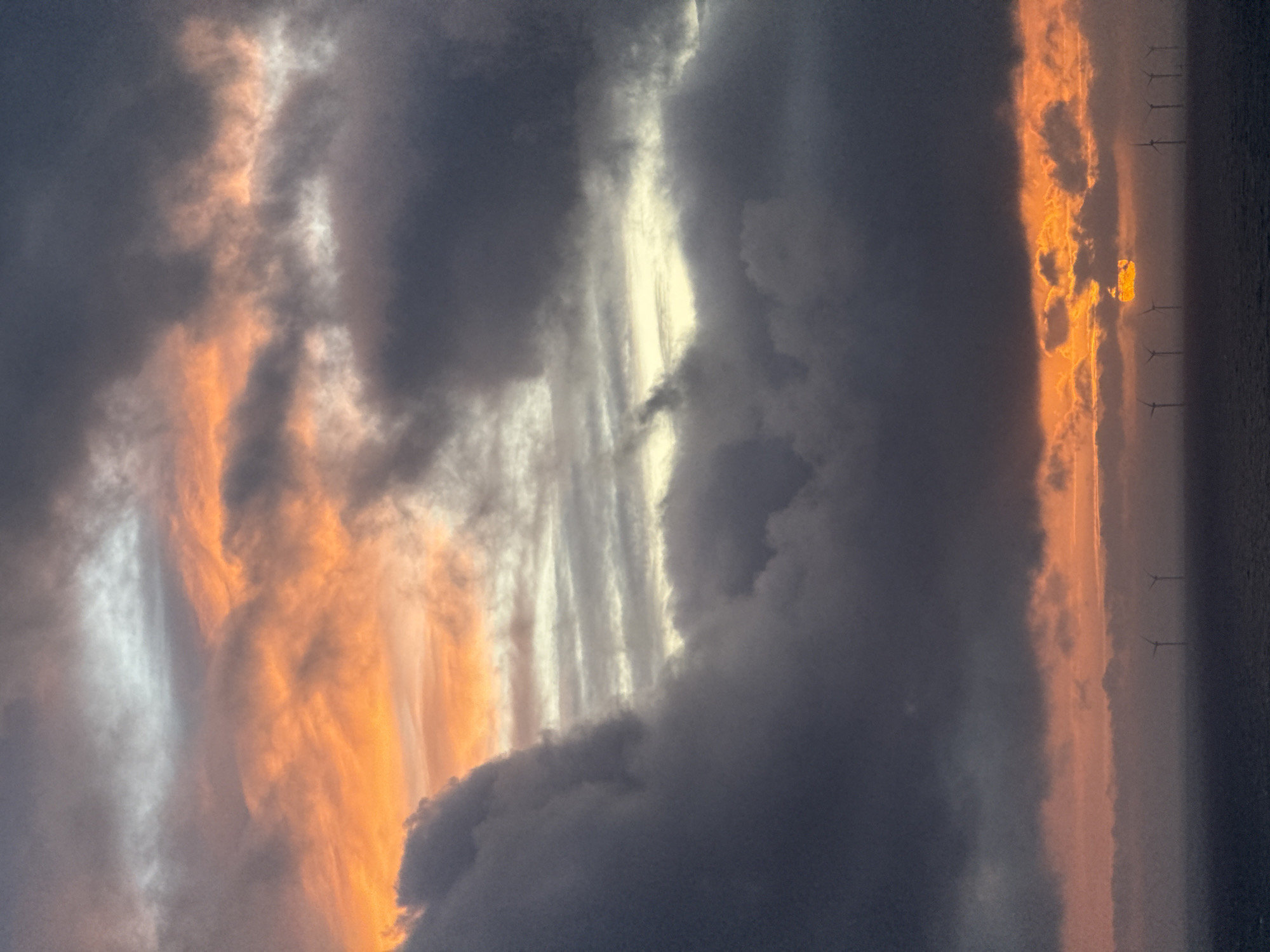 This media file captures a dramatic sunset or sunrise over a body of water, likely the sea, featuring a vast and expressive sky. The scene is dominated by a complex interplay of clouds, illuminated by the low-lying sun.
Large areas of the sky are filled with dark, heavy grey and bluish clouds, indicating a cloudy or potentially stormy atmosphere. These darker masses are interspersed with striking bands of light. In the upper-middle section of the sky, a prominent, elongated streak of bright, almost white-yellow light cuts horizontally across the frame, suggesting the sun's direct light piercing through or reflecting off higher clouds. Below this, closer to the horizon, another broad band of warm, intense orange and golden light illuminates the undersides and edges of numerous clouds, creating fiery textures and contrasts. Some clouds exhibit vertical streaking, catching the light in parallel lines.
Along the very low horizon, a series of dark silhouettes are visible against the orange glow: numerous offshore wind turbines, stretching across the distant seascape. This detail, combined with the location context, strongly suggests an offshore wind farm, possibly near Fécamp, France. The surface of the water beneath the turbines appears dark and calm.
No people are visible in the image, and there is no discernible text. The time of day is clearly the golden hour, and the overall mood is one of majestic natural beauty and powerful, dynamic weather conditions.