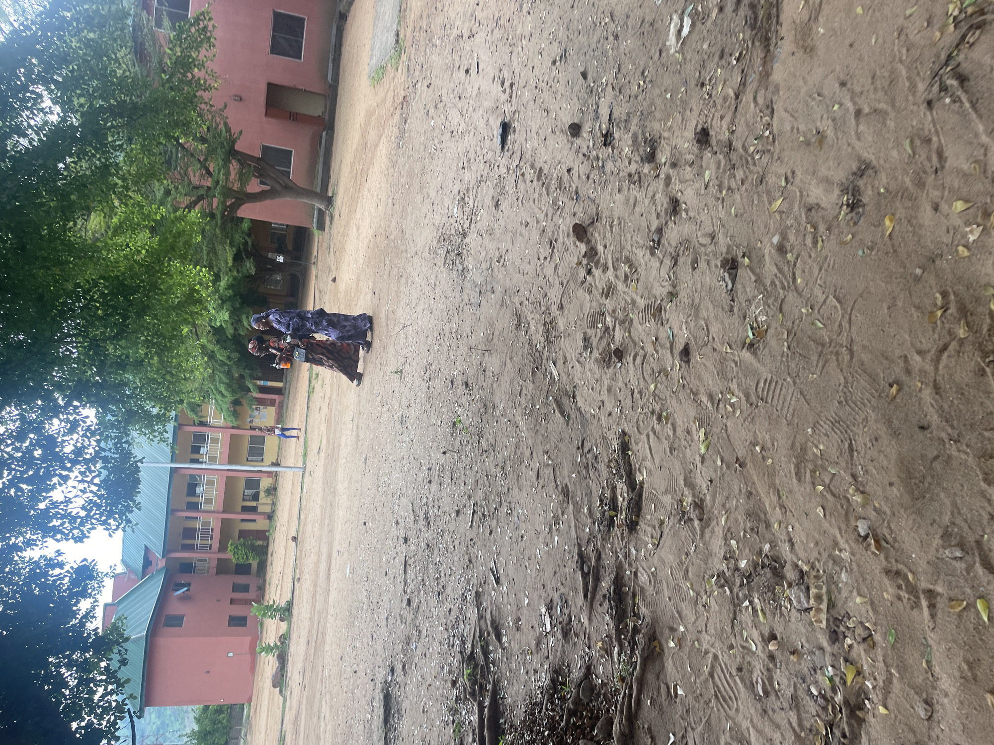 Two women in traditional attire are walking across a sandy, unpaved area in front of a pink and yellow multi-story building with a green roof. The building appears to be a school or administrative facility, with visible windows and balconies. The ground is covered in sand, scattered leaves, and some exposed tree roots. There are also footprints visible in the sand. In the background, more trees and another pink building can be seen. The scene suggests an outdoor environment within an urban or semi-urban setting, likely in a tropical or sub-tropical region given the foliage. The overall impression is one of casual movement and everyday life.