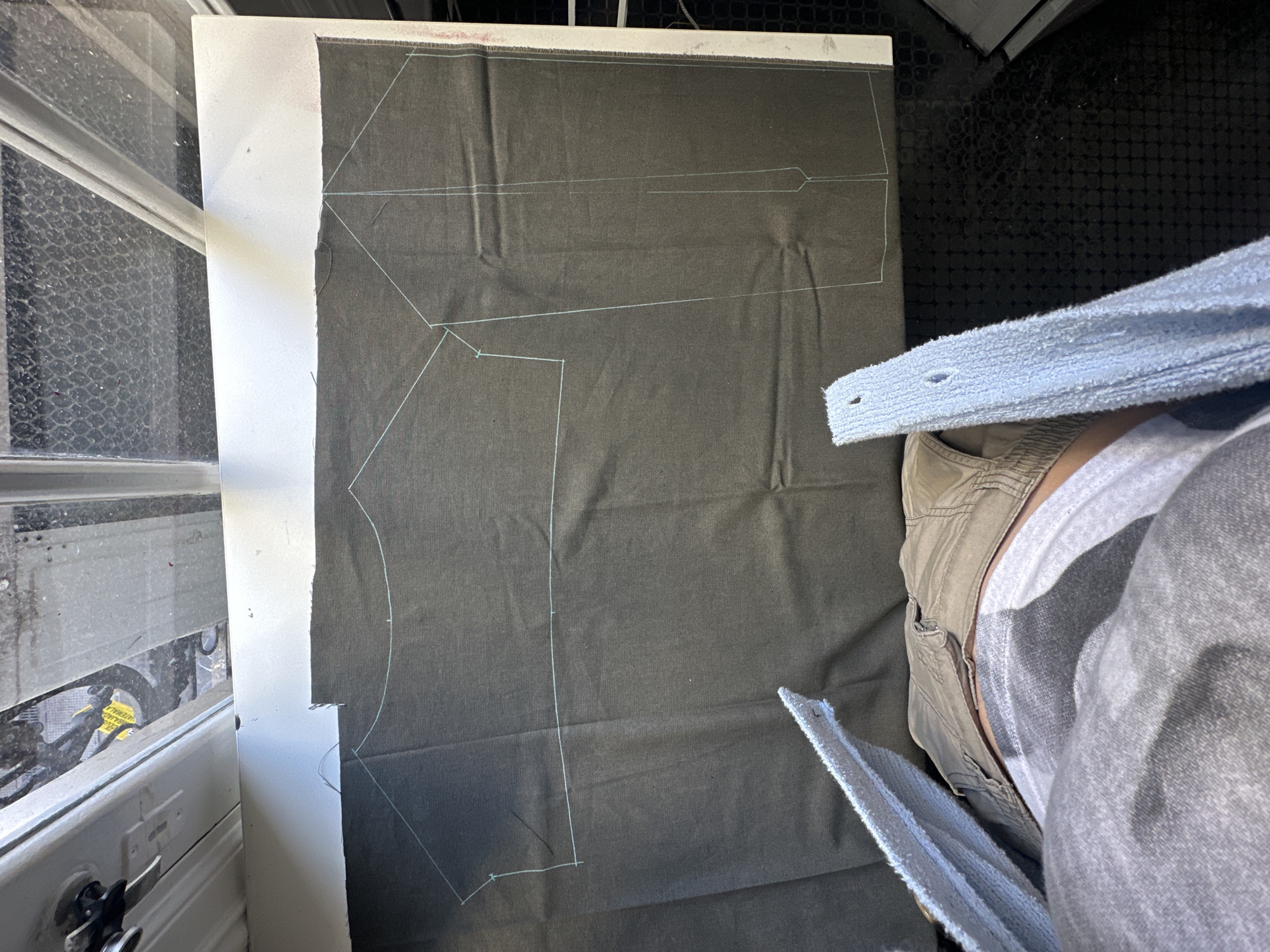 The image shows a section of dark fabric with a chalk outline of a pattern piece laid out on top of a piece of white cardboard. The fabric and cardboard are on a flat surface, possibly a table. A person is seated to the right, wearing a light-colored t-shirt and cargo pants, with a light blue towel draped over their lap. A window is visible to the left with a white frame and dirty glass. The environment is indoors, possibly a sewing or craft room. It's not possible to determine the location within Buenos Aires, Argentina, from the provided visual information.