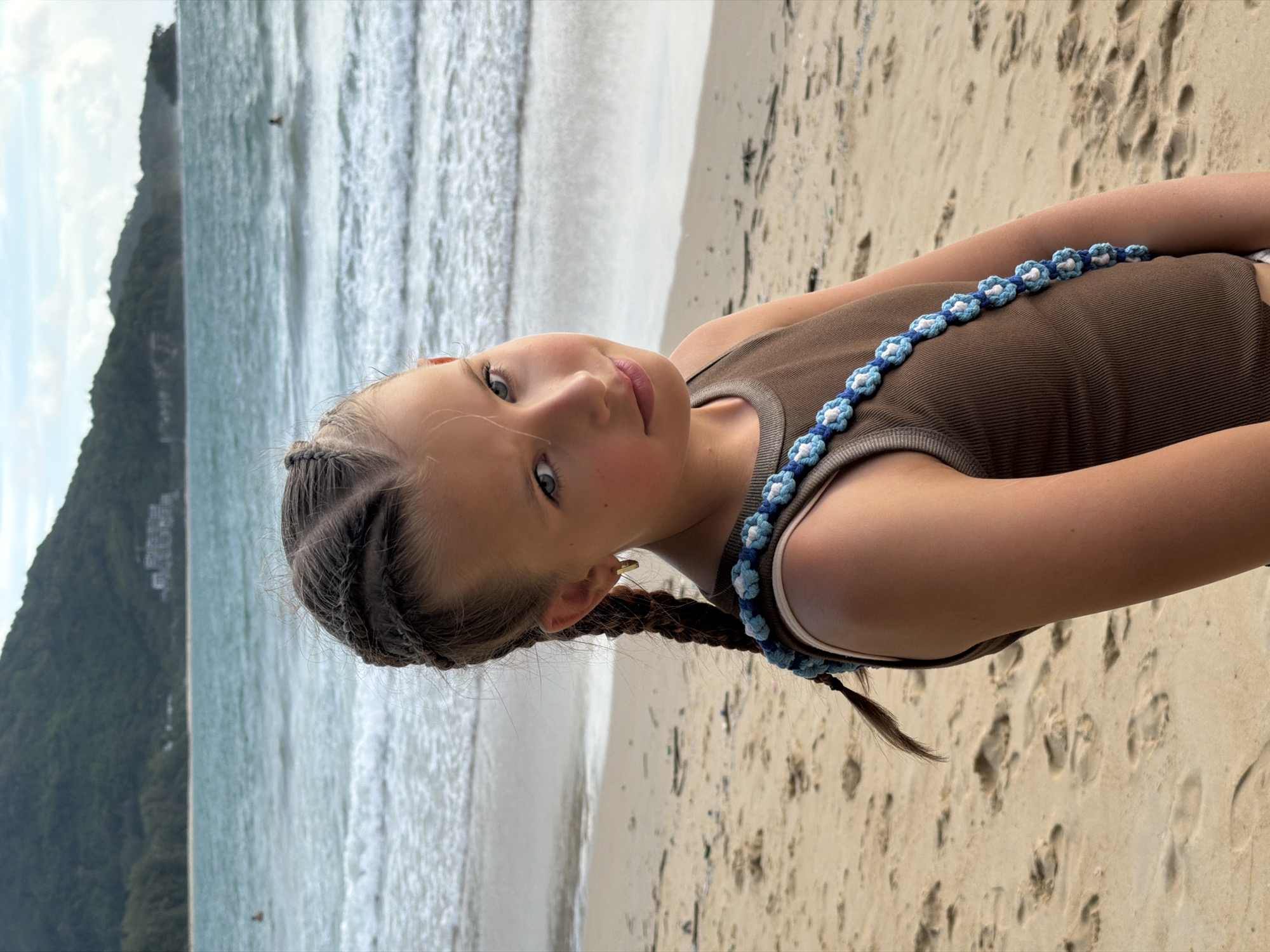 A young girl with braided hair and blue eyes stands on a sandy beach, facing the viewer. She is wearing a brown tank top adorned with a blue and white beaded strap across her chest. Her left ear has a gold hoop earring. In the background, the ocean stretches out with gentle waves, and a lush green, tree-covered mountain range rises in the distance. A few buildings are visible nestled on the mountainside. The sky is partly cloudy. The location is identified as Sakhu, Thailand.