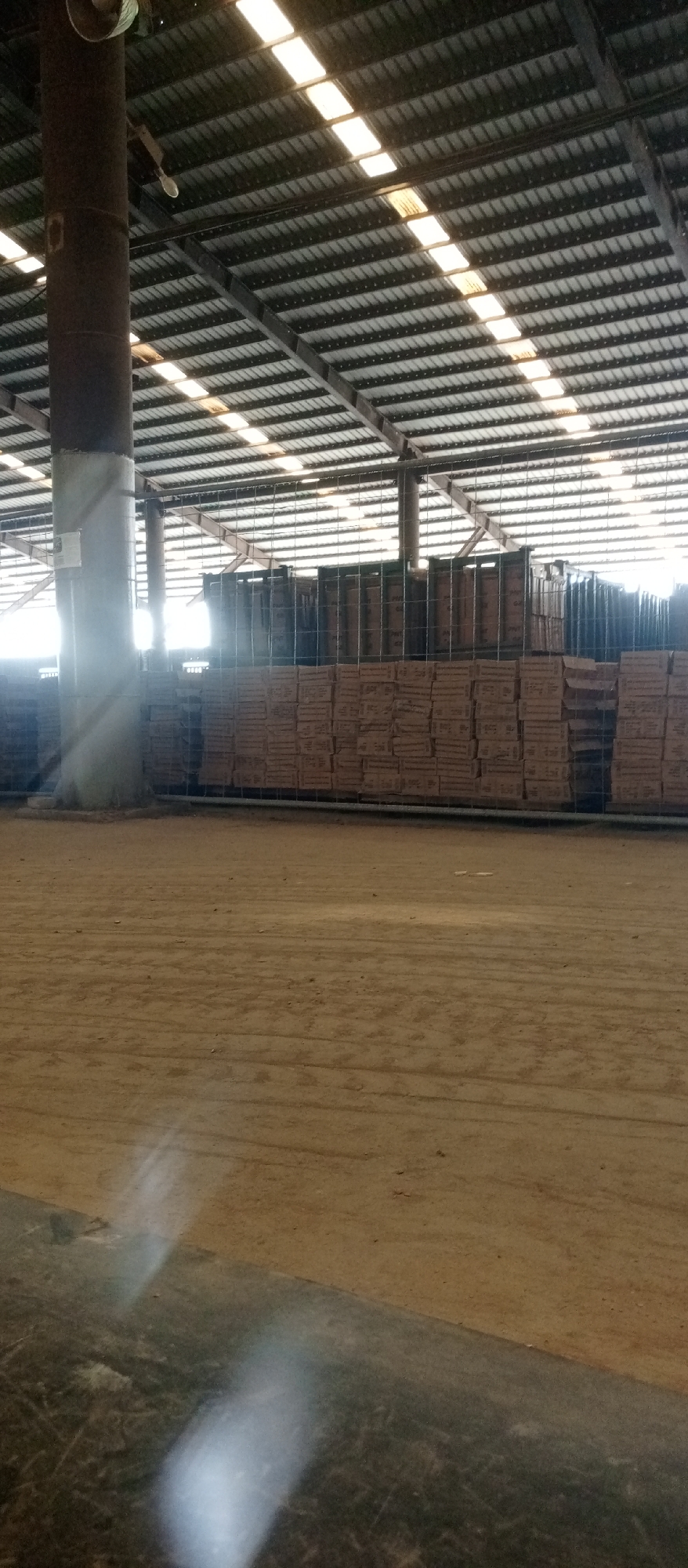 The image shows the interior of a large warehouse in Magboro, Nigeria. The warehouse has a high ceiling made of corrugated metal with translucent panels for lighting. A thick, cylindrical support column stands in the foreground, partially obscuring a stack of boxed goods behind a wire mesh fence. The floor is dirt or packed earth. There are no people visible in the image. The light is bright, with some glare.