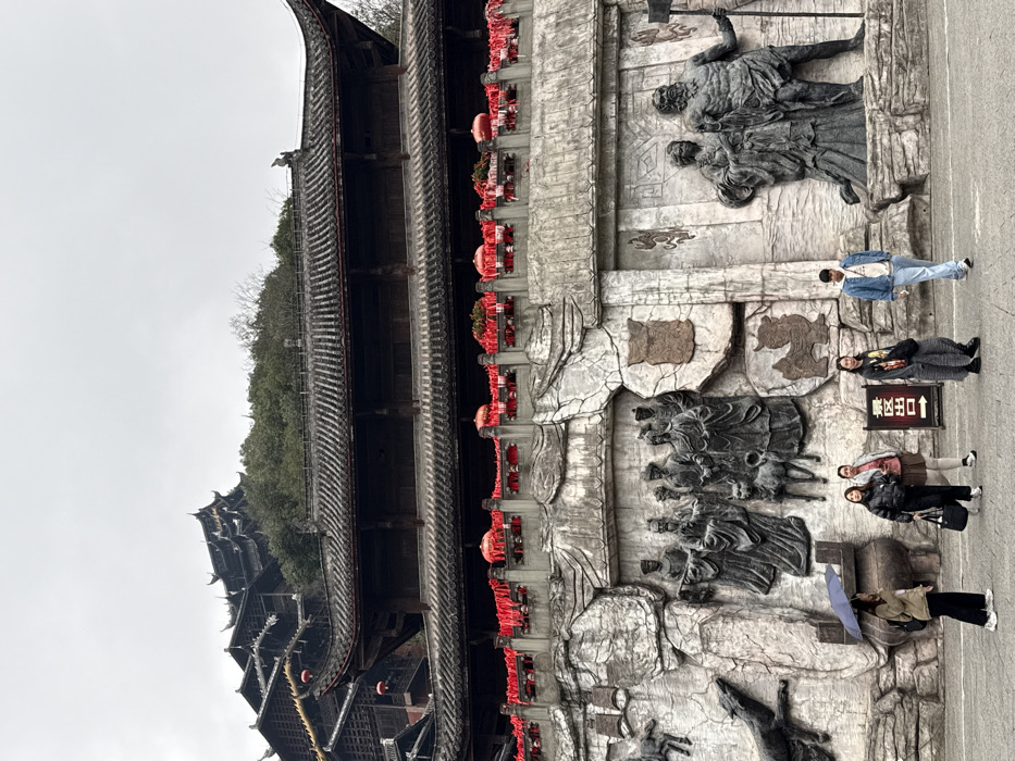 The image displays a traditional Chinese architectural complex in Pengshui Miao and Tujia Autonomous County, China, under an overcast sky. Multiple dark-roofed buildings, characterized by their tiered structure and upturned eaves, ascend a tree-covered hillside in the background. In the midground, a large building with an ornate dark tiled roof features a lower-level balcony adorned with numerous red lanterns and red ribbons strung along its railing. Beneath this structure, a prominent stone wall displays extensive bas-reliefs depicting human figures in traditional attire, various animals, and symbolic elements, rendered in a grey, textured stone. The foreground consists of a paved ground area. Six individuals are visible: a woman on the far left holds a black umbrella; a woman in a black puffer jacket and dark pants, carrying a black tote bag, stands next to a woman in a light-colored top and a beige skirt; a fourth woman in a dark patterned long-sleeved shirt and grey plaid pants stands near a black and white sign marked "出口" (Exit) in Chinese characters; a man in a blue denim jacket and light blue jeans walks rightward across the frame; and another person is partially obscured behind the man in the denim jacket.