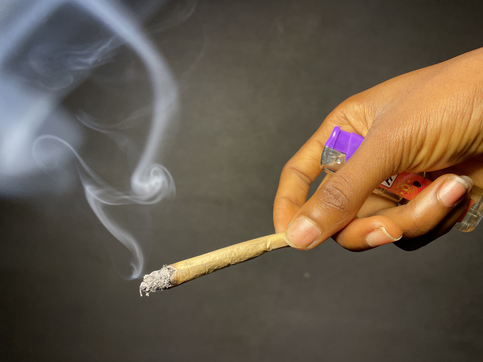 A tan hand holds a lit, rolled cigarette with ash on the lit end. Smoke rises from the cigarette. A purple and clear lighter is also held in the hand. The background is black. The location is not determinable based on the image.
