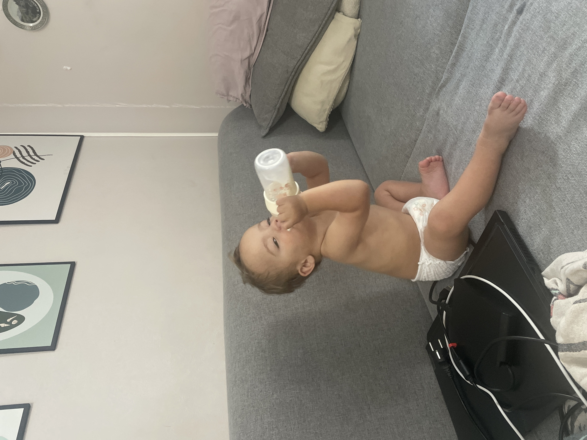 A toddler wearing a diaper is sitting on a gray couch and drinking from a bottle. The toddler is facing away from the camera and is holding the bottle with both hands. The couch is in a living room, and there are several pillows on the couch behind the toddler. There is also a laptop on the couch in front of the toddler. The walls are painted a light color and there are several framed pictures on the wall. The scene is set in Almaty, Kazakhstan.