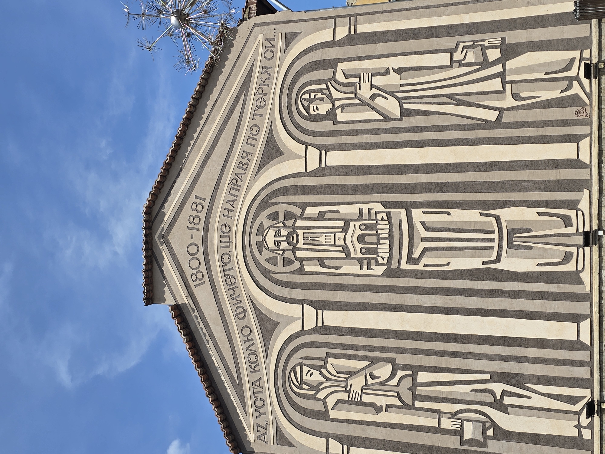 A low-angle, daytime shot focuses on a building facade. The facade features horizontal bands of beige and gray. Three arched alcoves are integrated into the design. Each alcove contains a stylized figure, rendered in beige and gray to match the building's palette. Above the alcoves, the facade is topped with a peaked roof adorned with dark brown tiles. Etched into the facade above the alcoves is text in a foreign language, along with the years "1800-1881". In the upper left corner, a metallic, starburst-like sculpture is visible against a backdrop of blue sky dotted with scattered clouds. The location of the building is Unknown, Unknown.