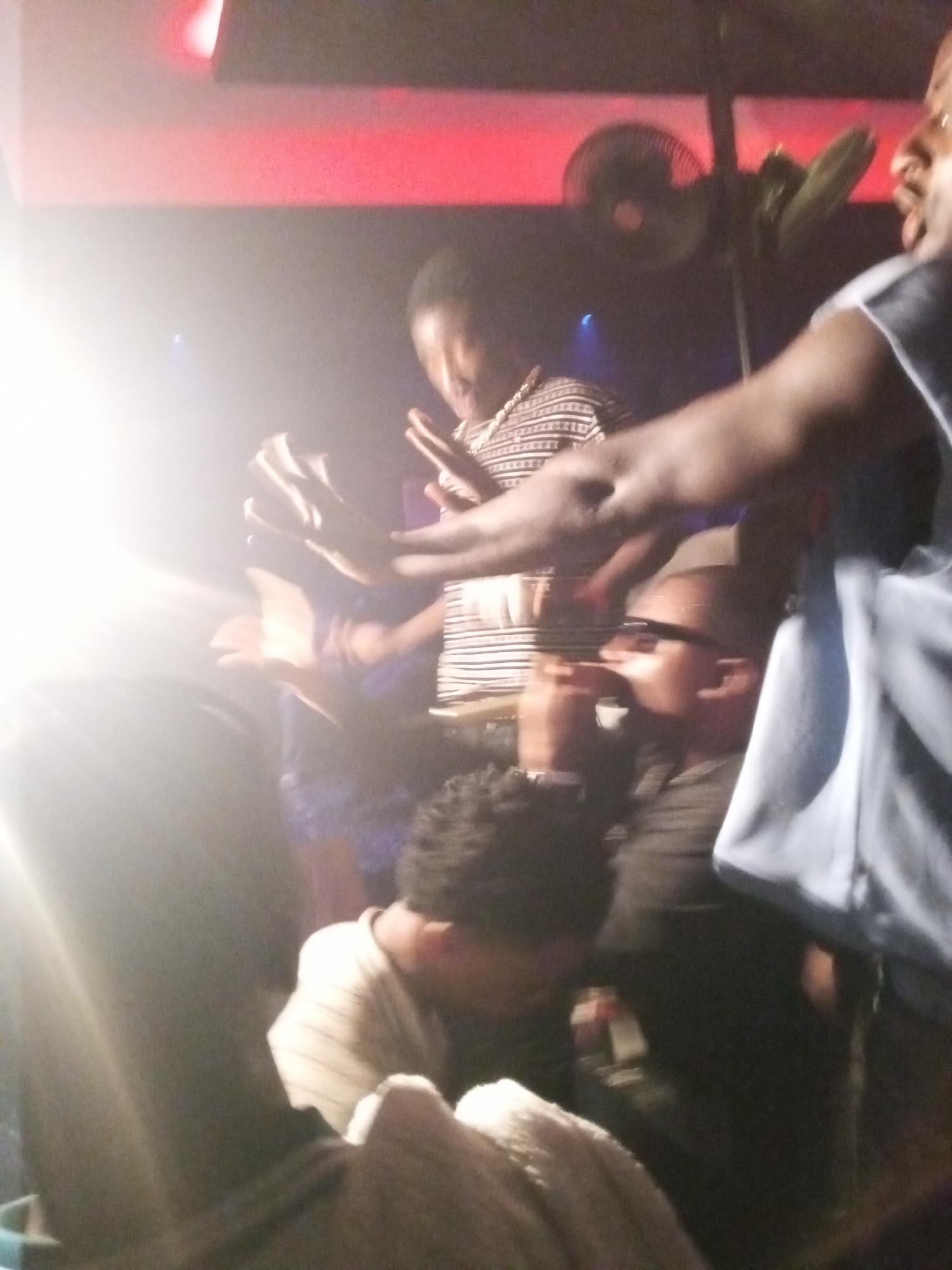 The image appears to be taken indoors, likely in a crowded and dimly lit setting, potentially a club or gathering place in Lokoja, Nigeria. Several people are visible, including one in a striped shirt raising their hands. Another person wears sunglasses. A fan is mounted in the background, and there is red lighting on the ceiling. The overall lighting is poor, with a bright glare obscuring parts of the image.